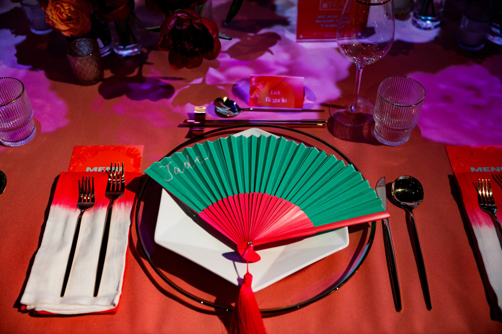 Green and red folding fan with a red tassel on a plate between black cutlery and orange menus