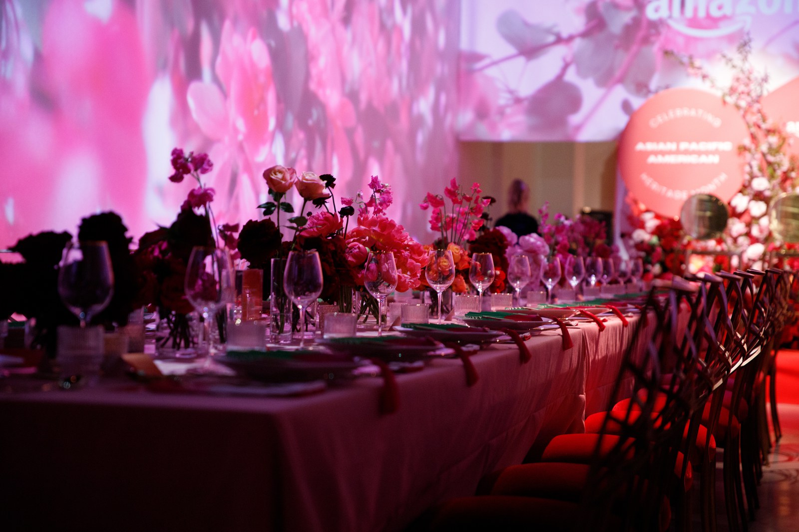 Long dinner table with pink and red flowers and glassware, an Amazon logo projected on the blossom lit wall behind