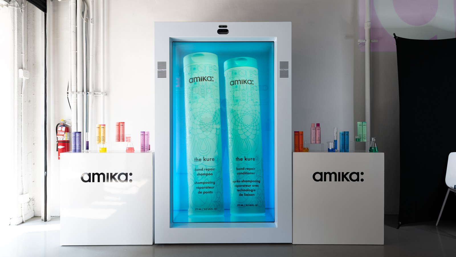 Two giant blue Amika bottles glow inside a lit white display case flanked by plinths of colourful products