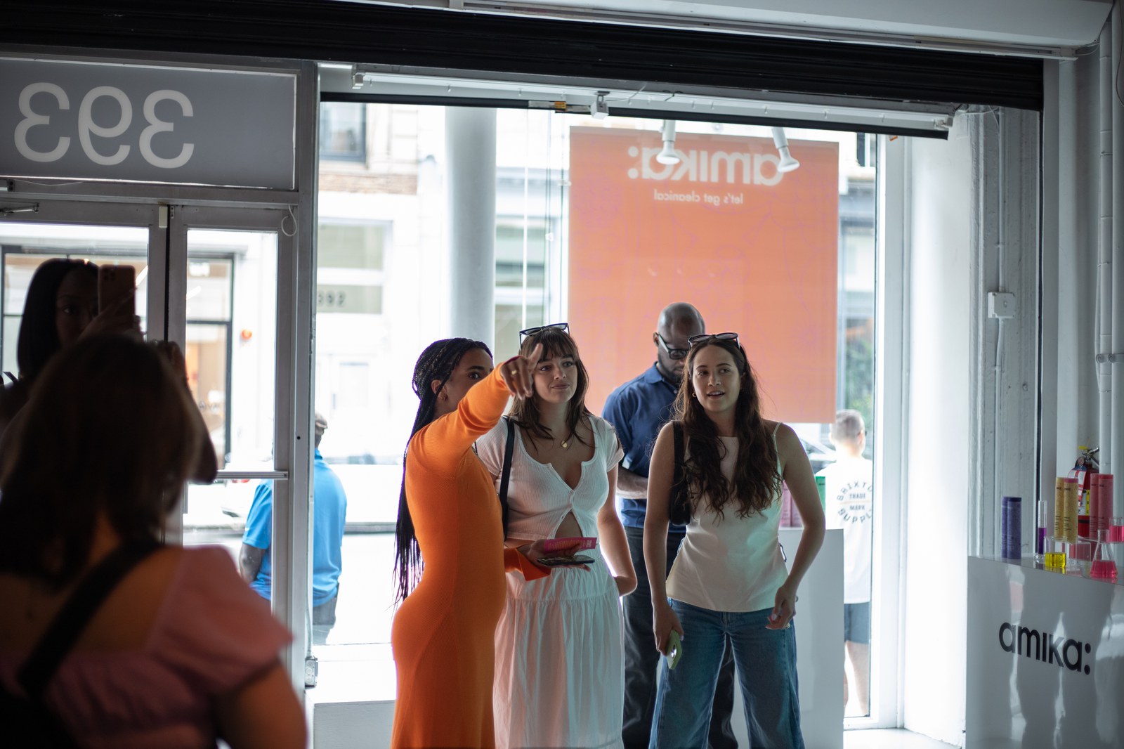 Guests in white and orange pause near the open street entrance of a bright Amika storefront