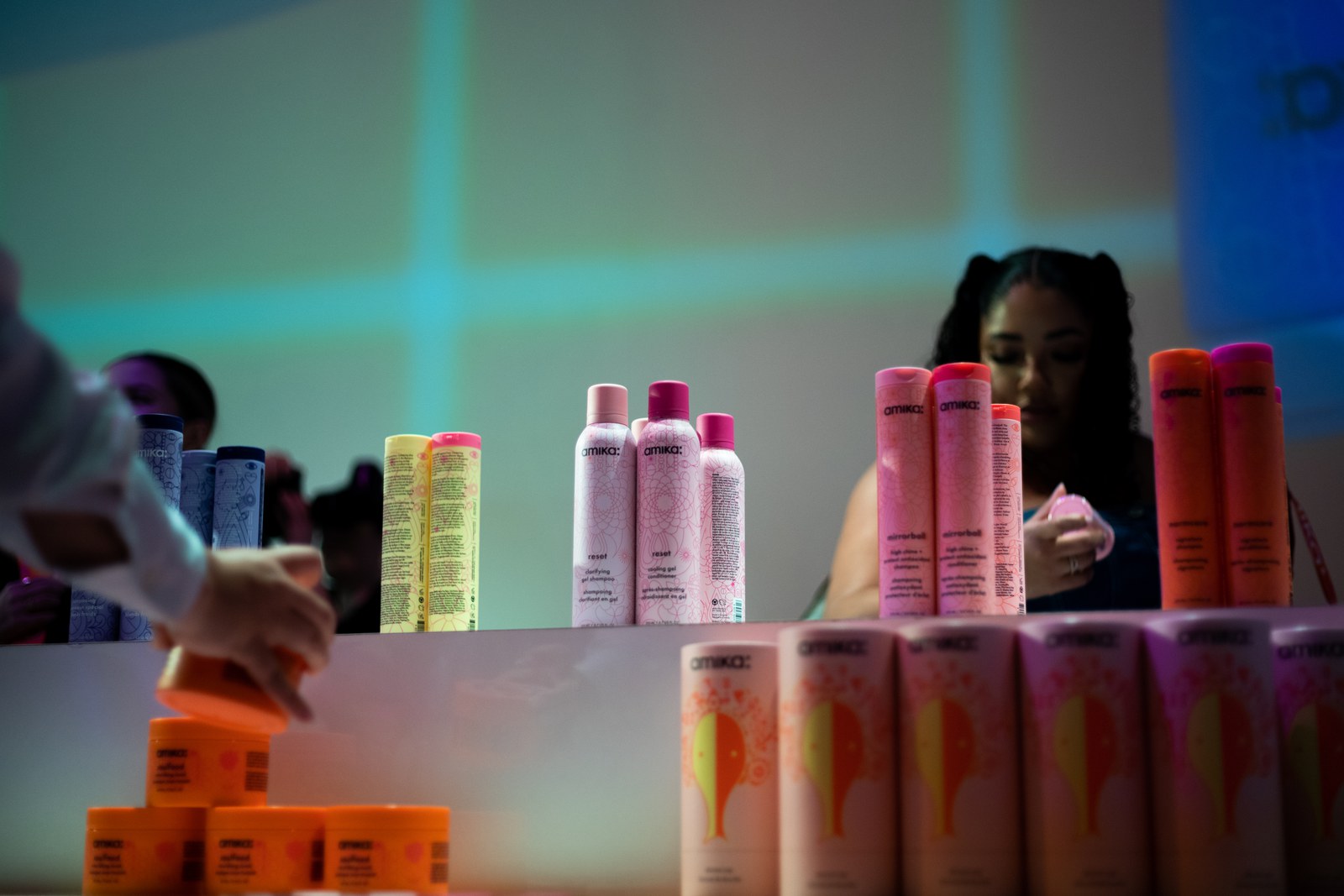 Pink, orange and yellow amika spray cans on a white counter with a hand reaching for a jar