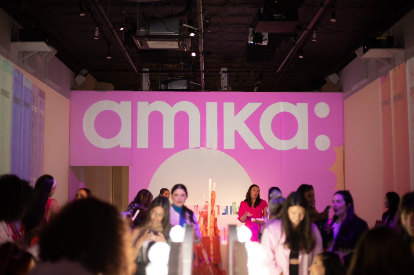 Guests gather in pink light beneath a large amika wall logo as two women stand at a lit product bar