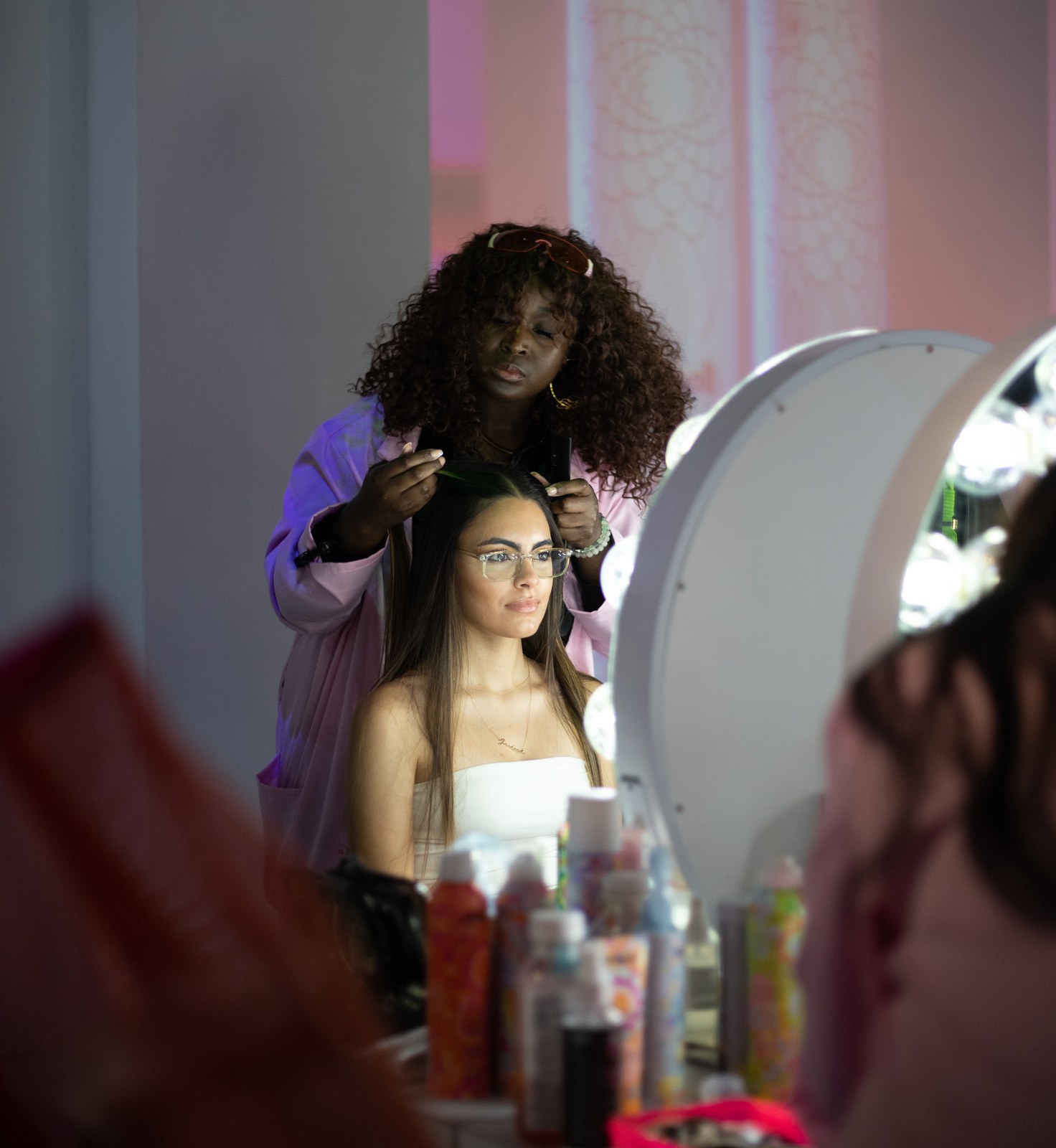 Hair stylist working on a seated client in white in front of a round bulb lit vanity mirror
