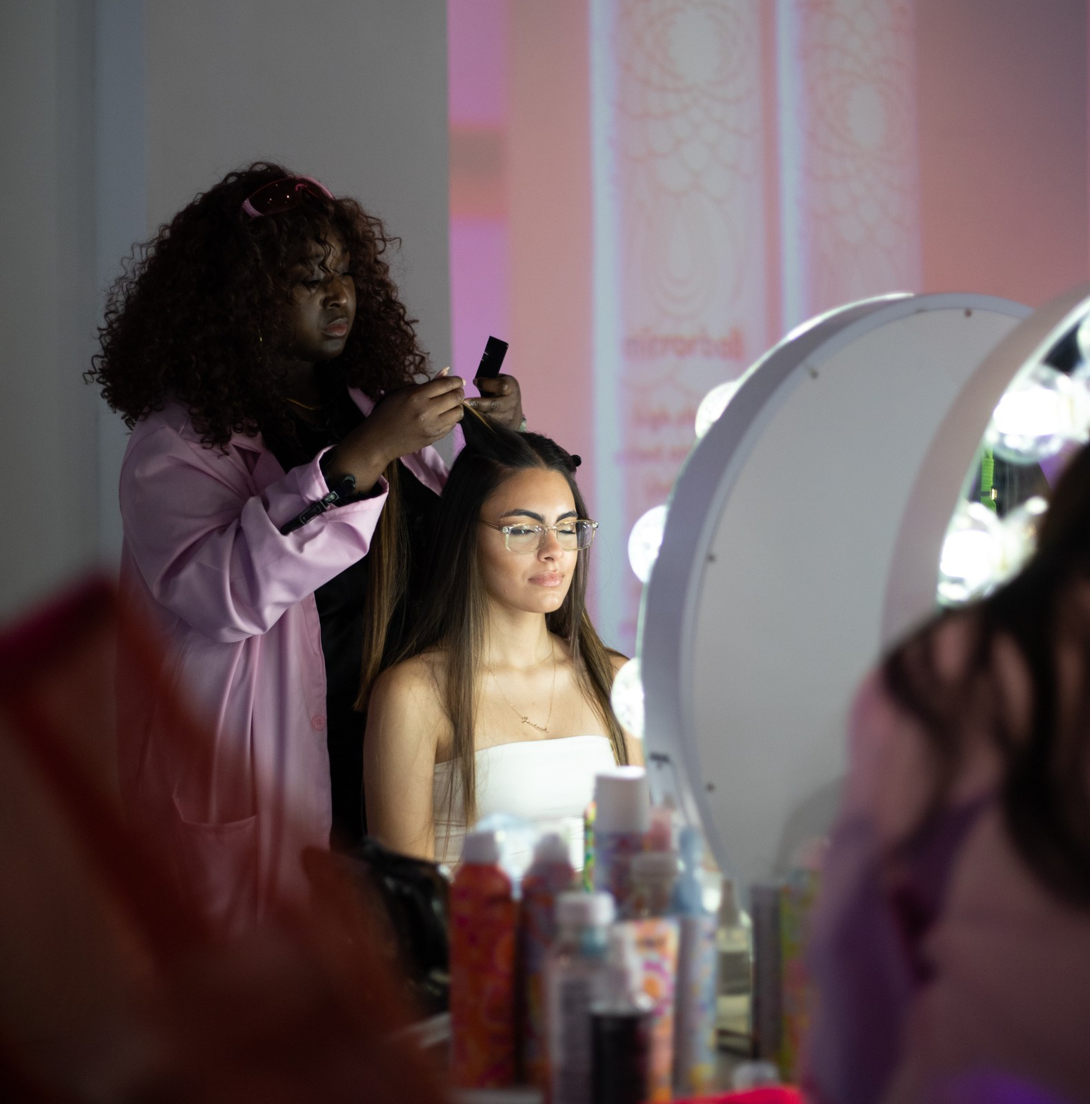 A stylist in a pink coat combs a seated guest’s hair beside a lighted mirror and bottles of product