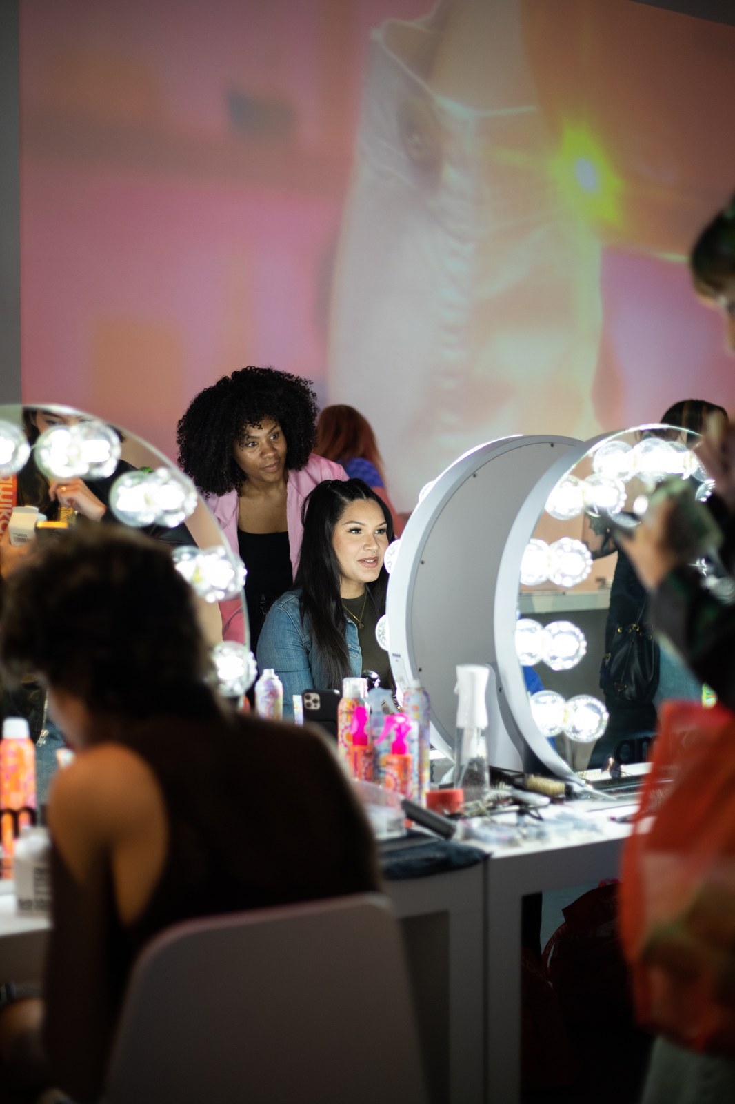 Two women sit at lighted round vanity mirrors surrounded by hair products, a pink projection glowing on the wall behind