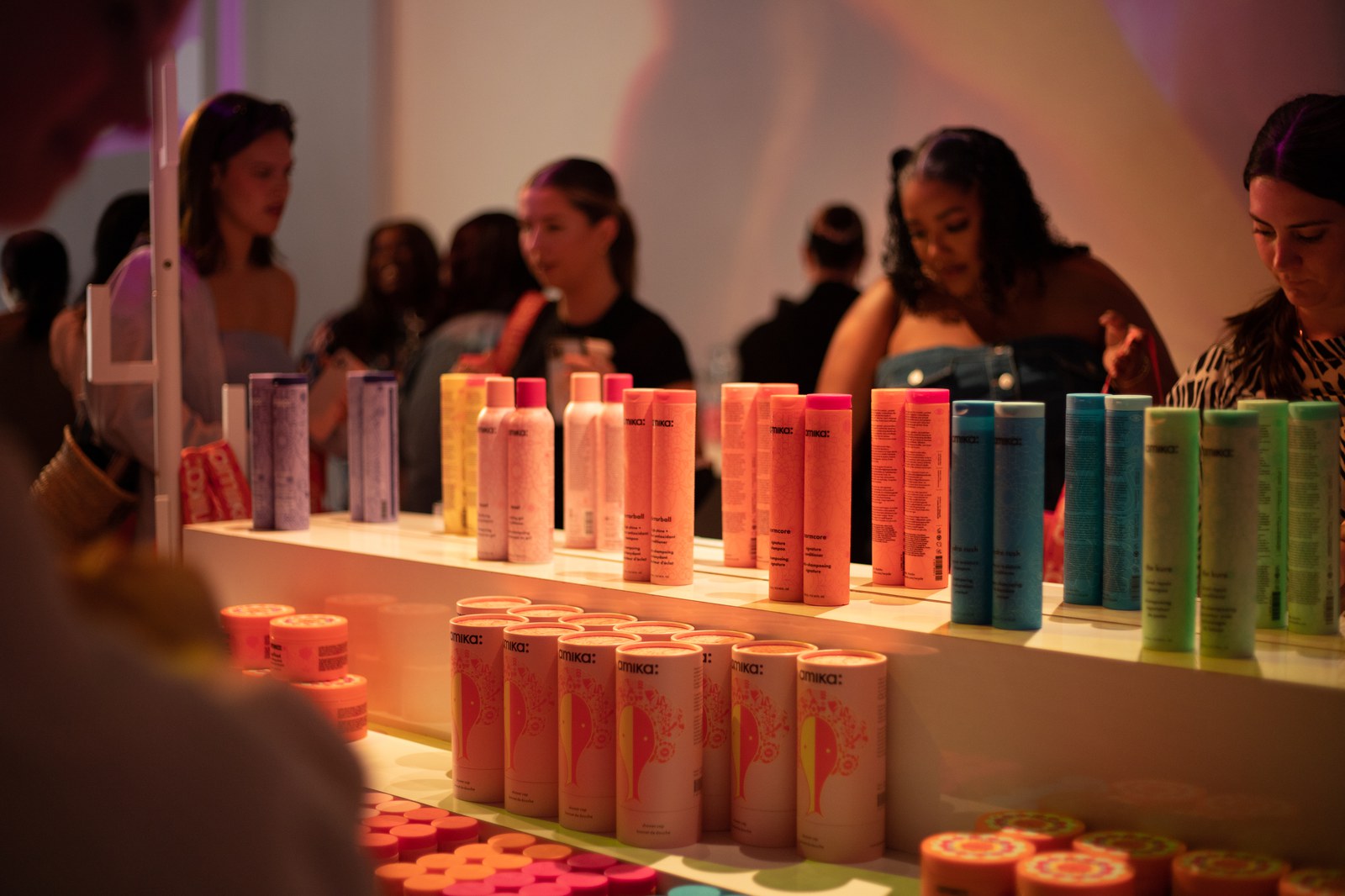Rows of pastel amika haircare bottles on a white shelf with guests browsing behind under warm light