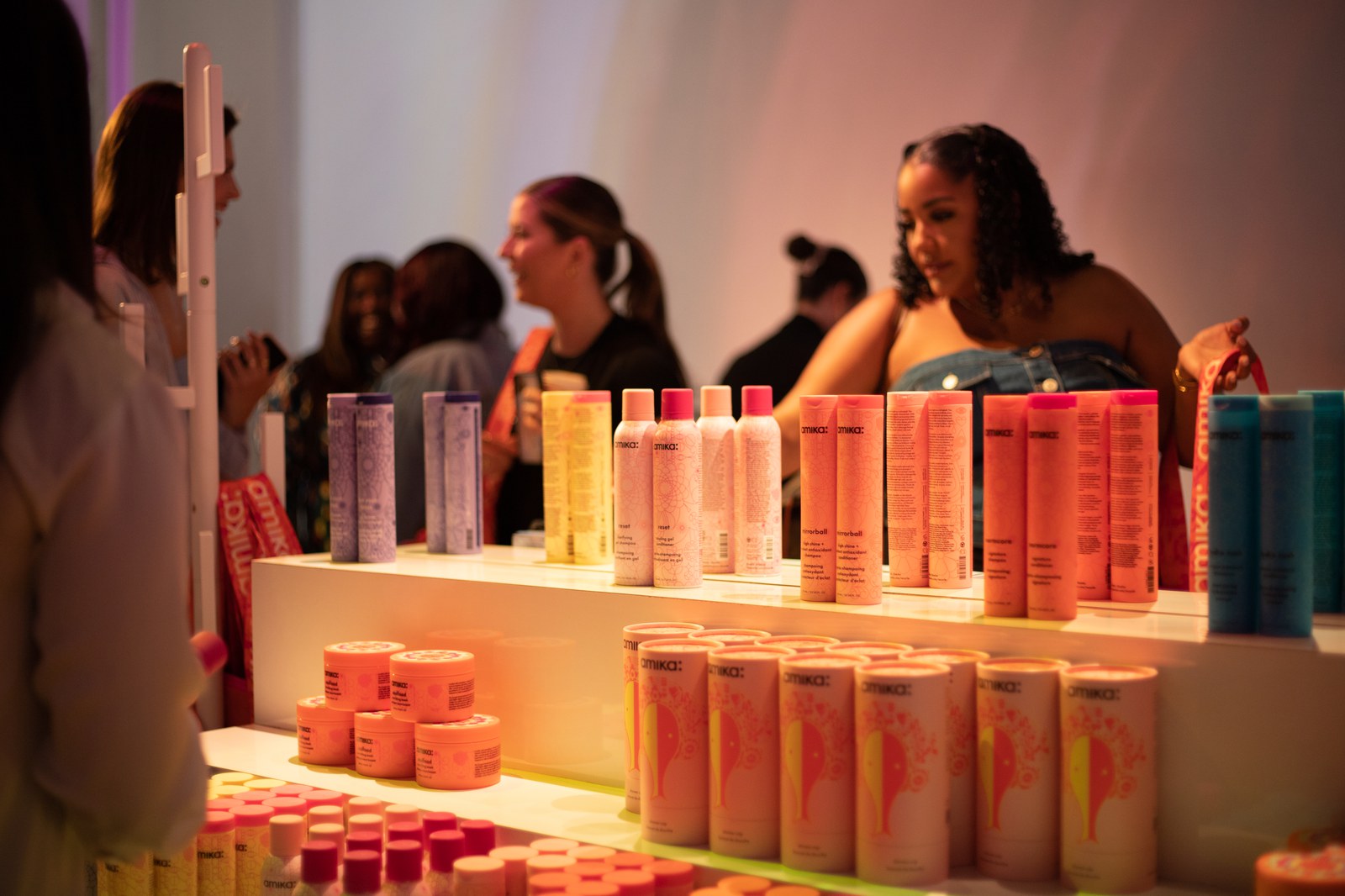 Rows of pink, peach and lilac Amika bottles and jars on white risers with guests chatting behind