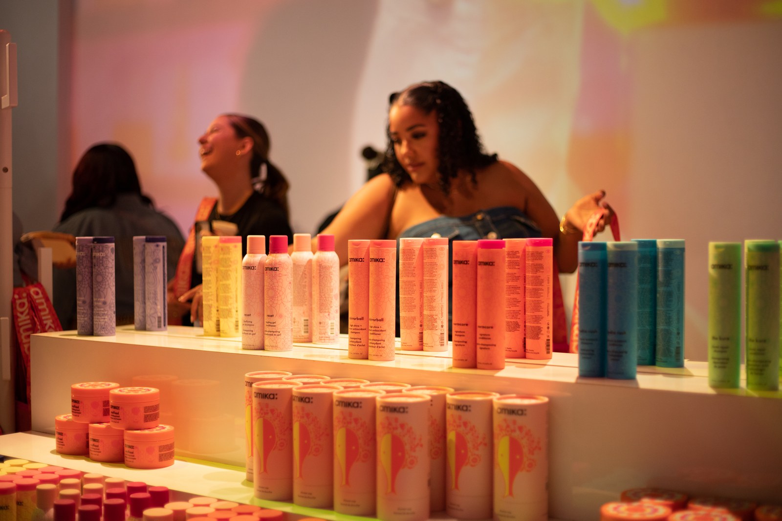 Rows of pastel amika hair product bottles and jars on white shelves with guests chatting behind them
