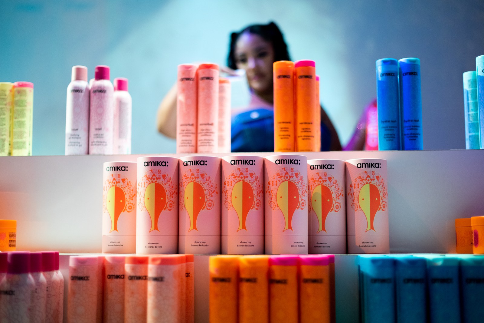 Peach and orange Amika cans stand in a row on a white shelf in front of blue and pink bottles