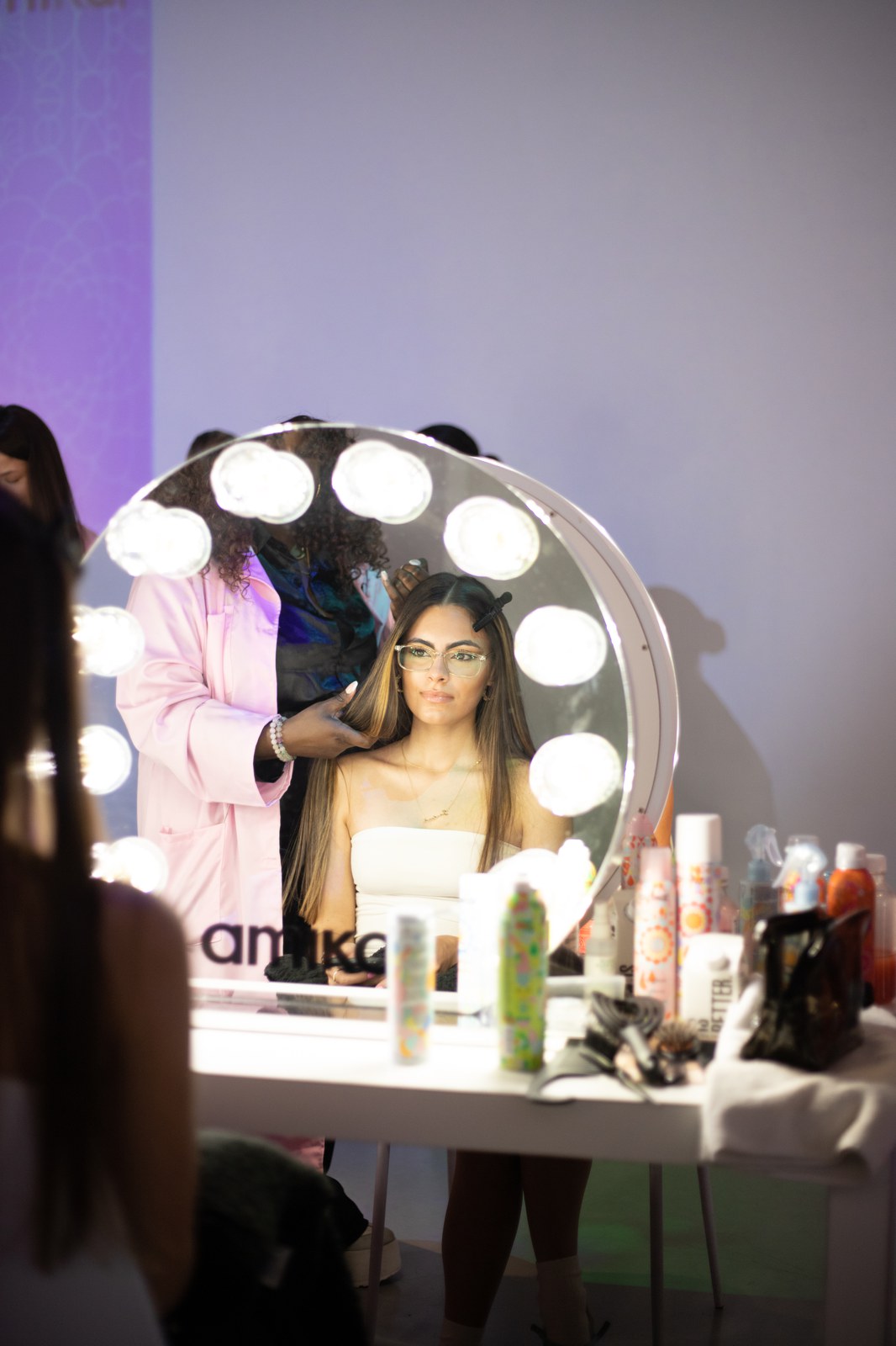 A seated woman in glasses is reflected in a round bulb lit mirror bearing the Amika logo