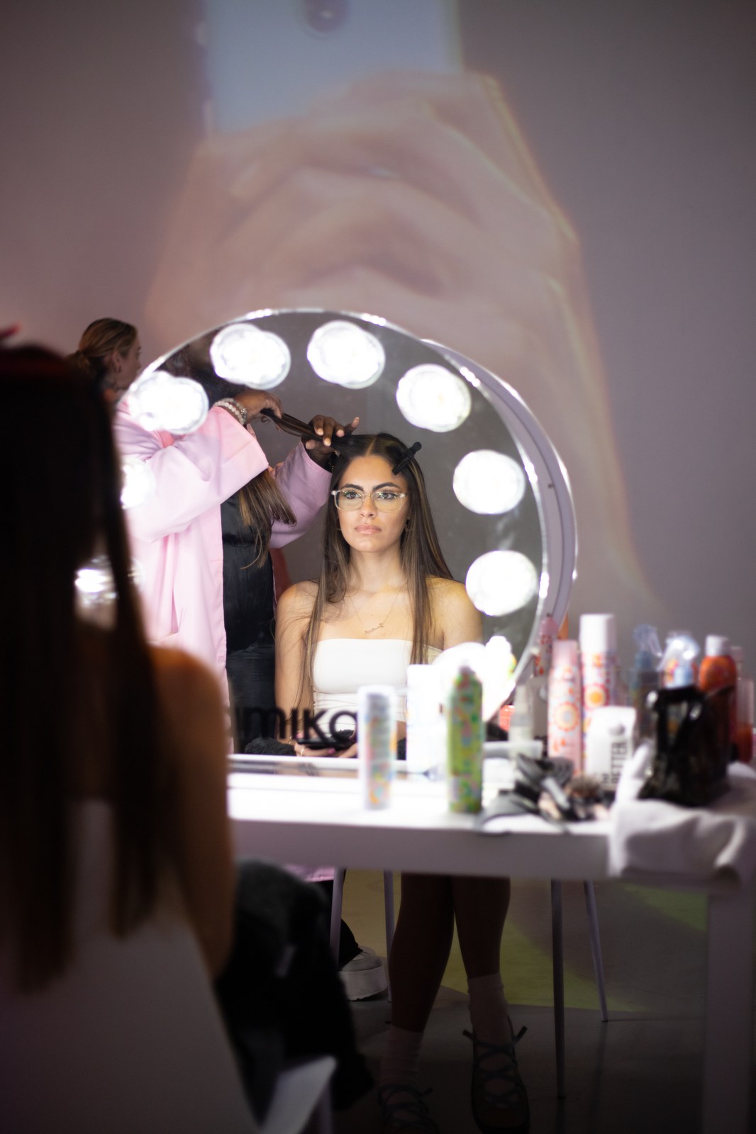 A stylist in pink curls a seated woman’s hair at a round lighted mirror marked with an amika logo