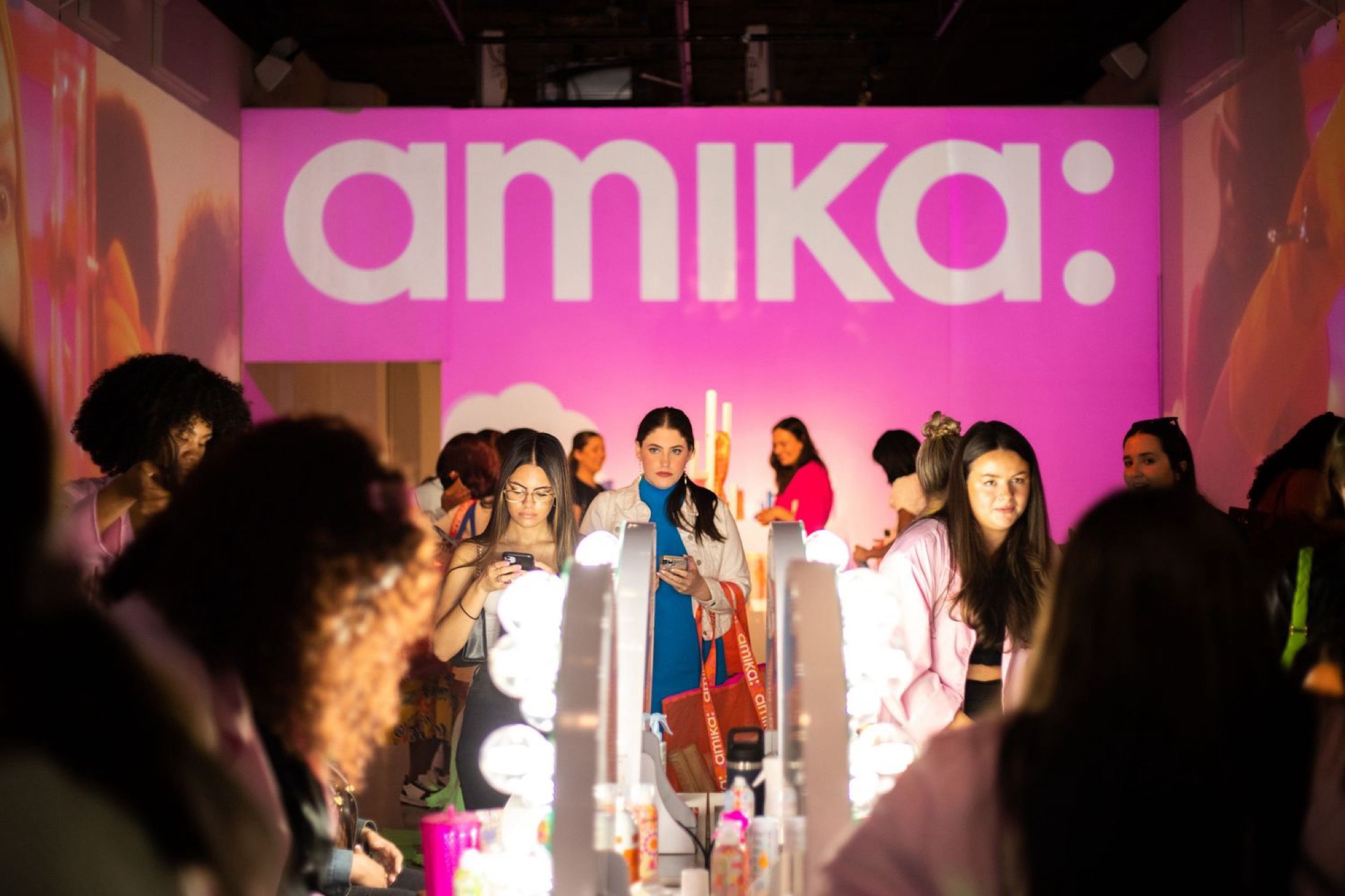 Guests gathered near lit vanity mirror stations under bright pink Amika branding at the fashion week activation