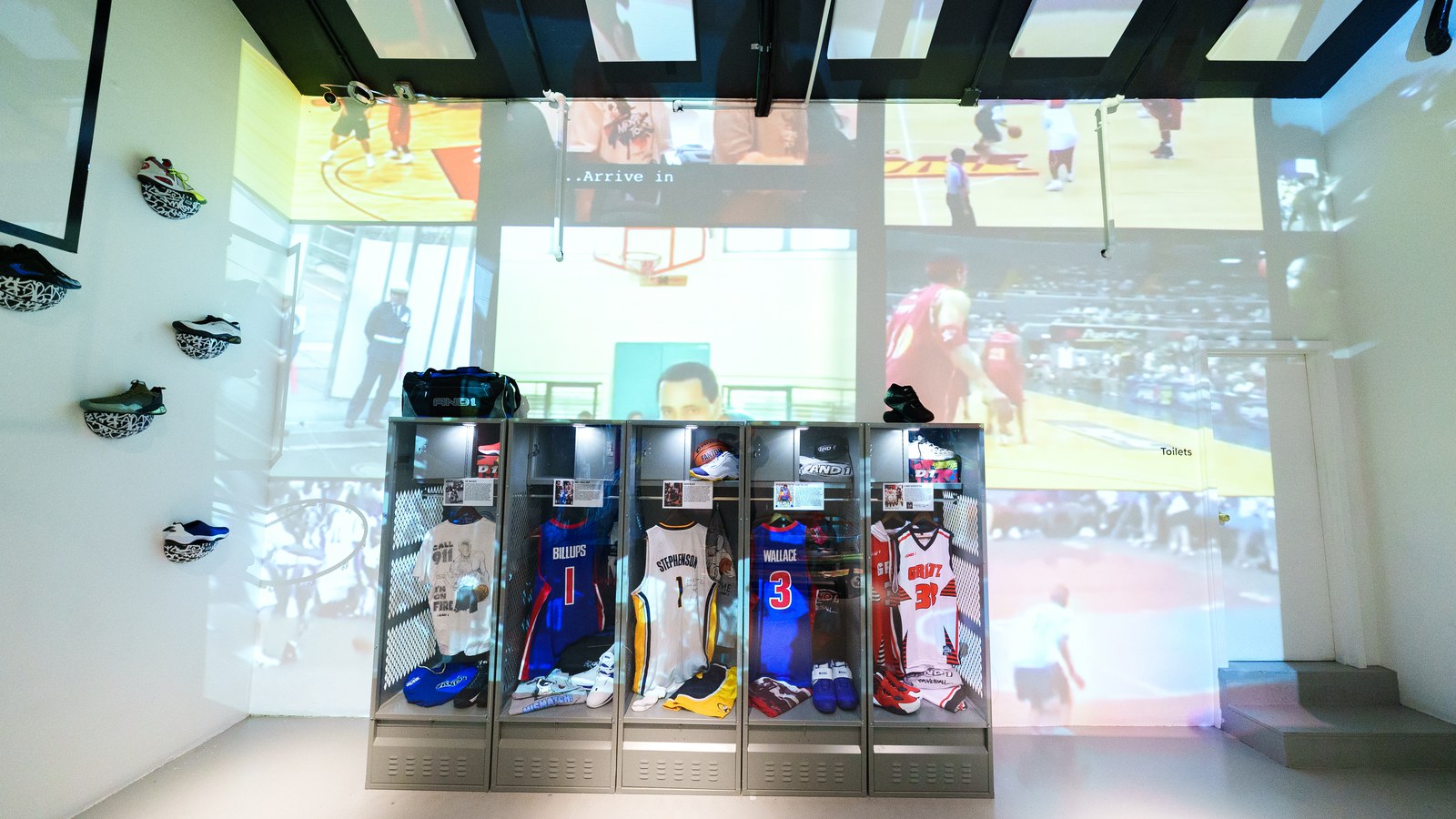 Glass locker cases holding basketball jerseys beneath projected game footage, with sneakers mounted on a white wall