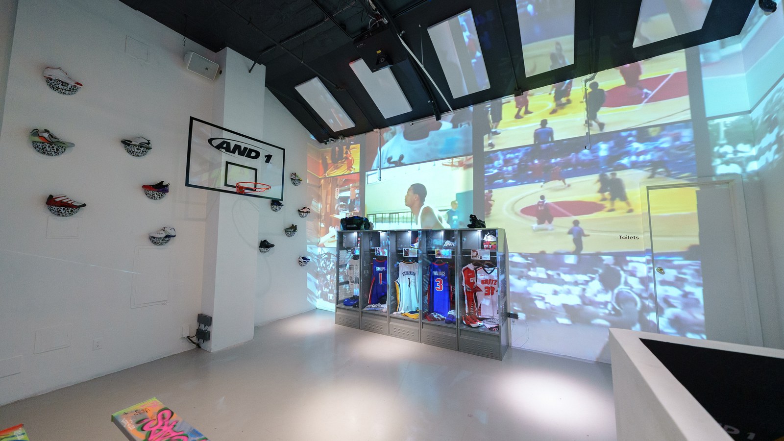 AND1 backboard and sneakers mounted on a white wall beside glass lockers of jerseys under projected game footage