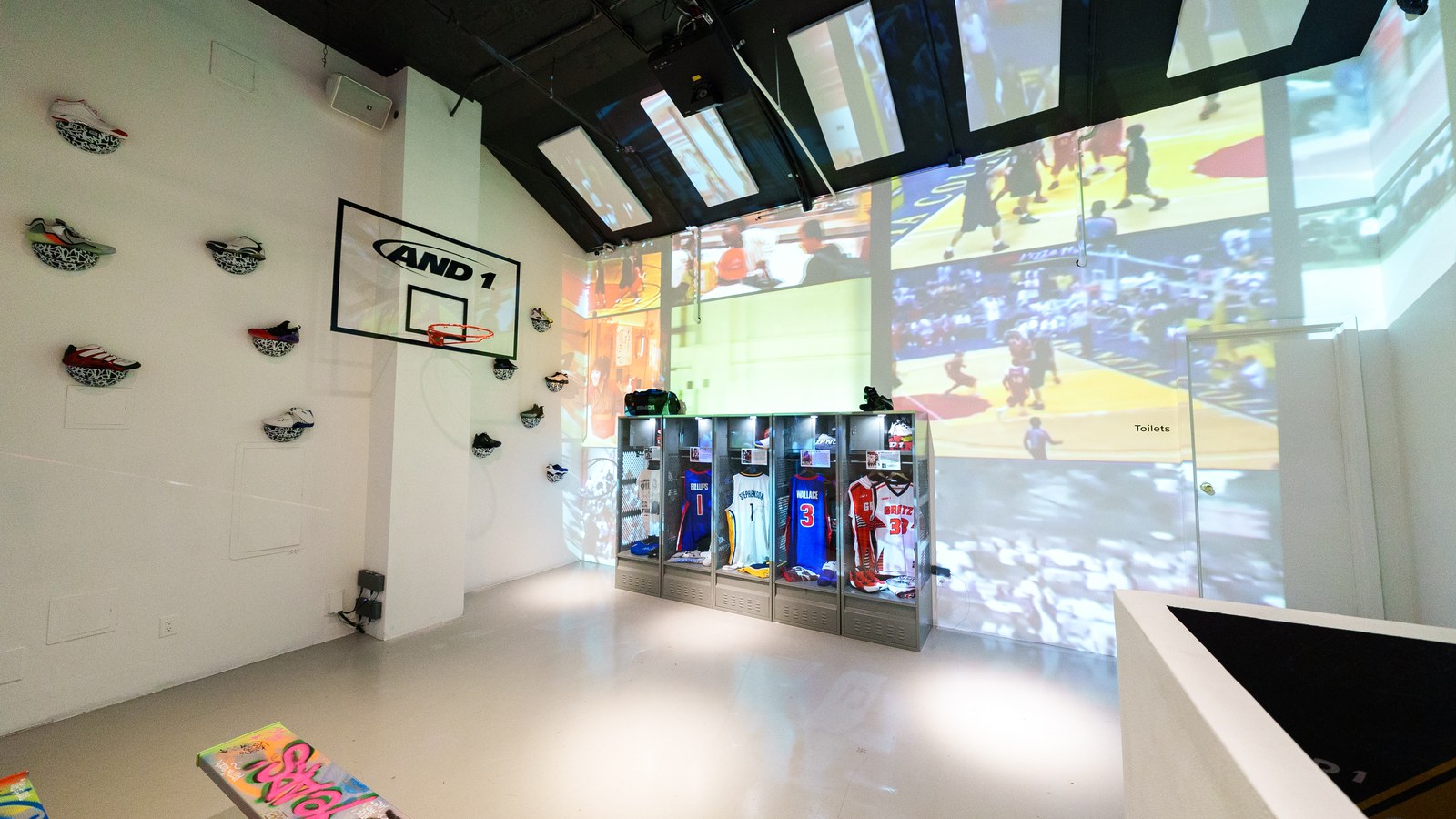 Wide view of a white showroom with an AND1 backboard, projected basketball footage and jersey lockers