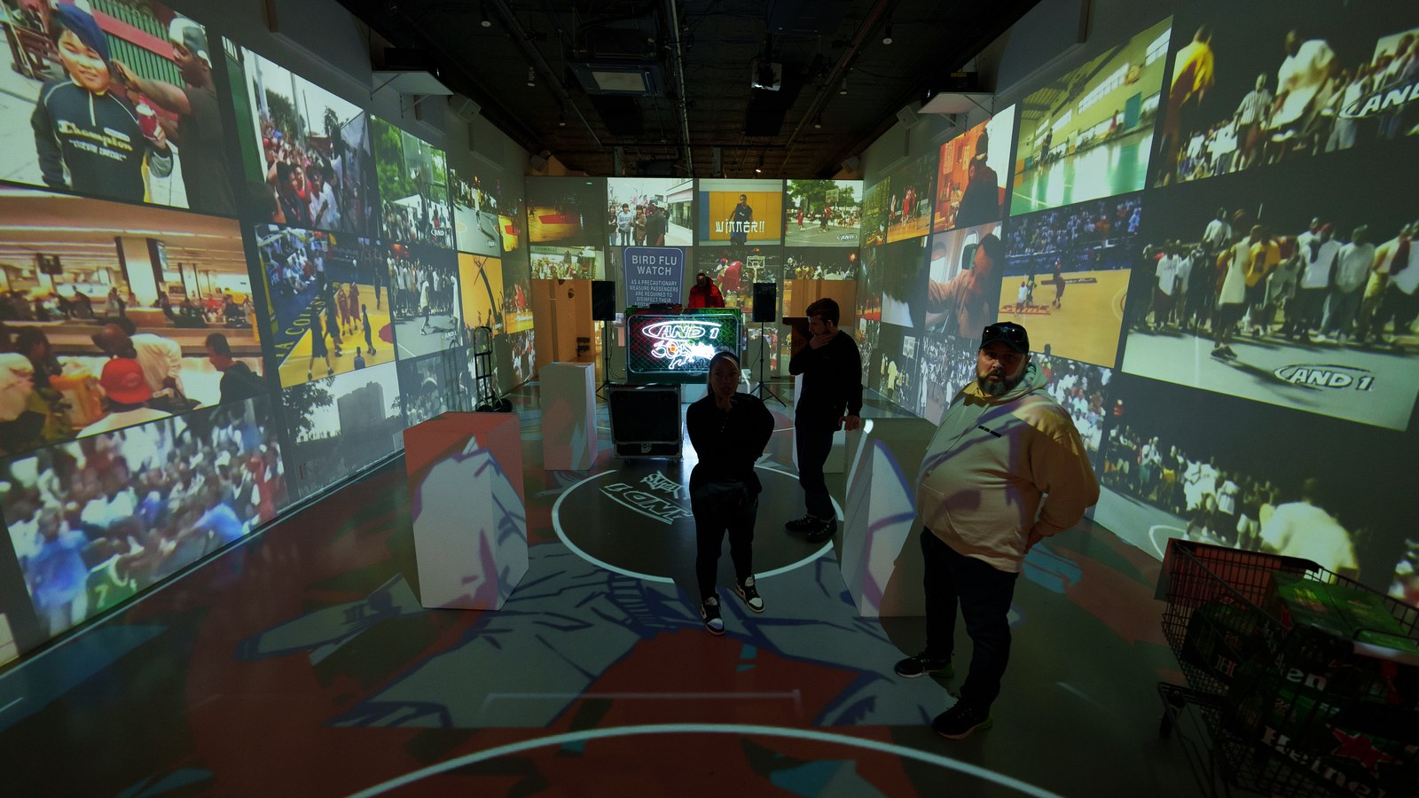 Three visitors stand on a painted court floor surrounded by walls projected with basketball crowd footage