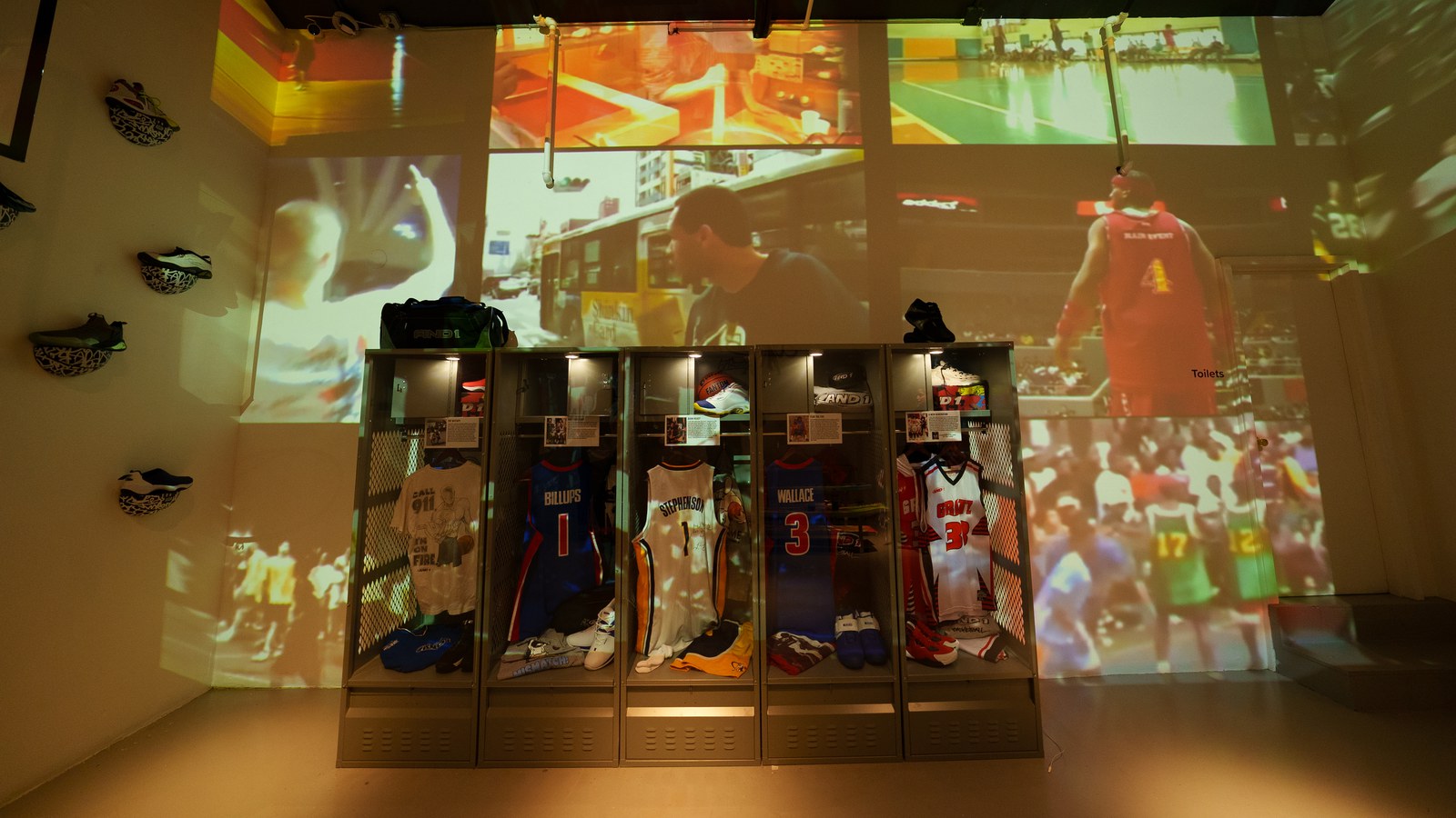 Basketball game footage projected across dim walls above open lockers filled with jerseys and sneakers