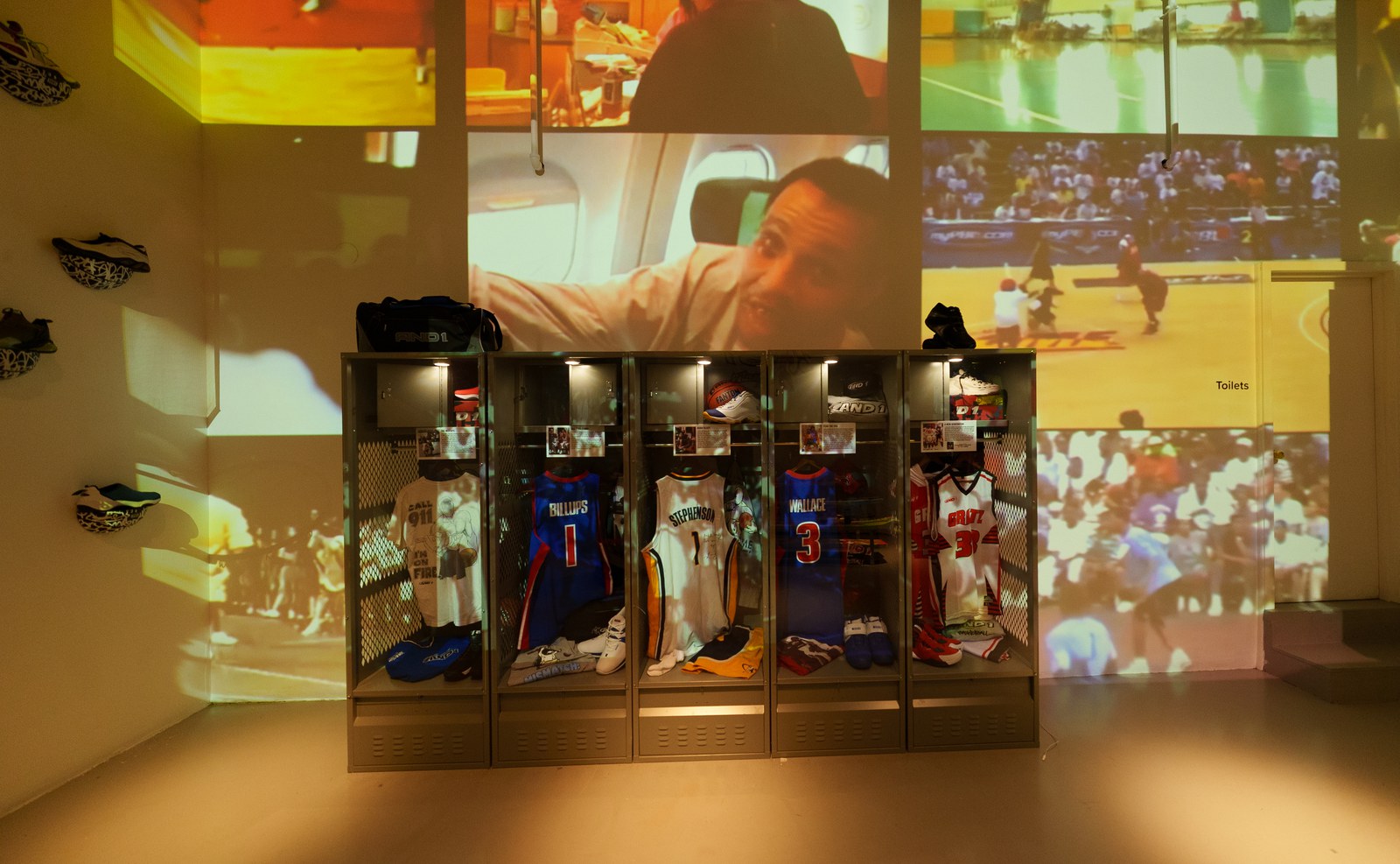 Five lit locker cases of numbered basketball jerseys in front of warm projected basketball footage