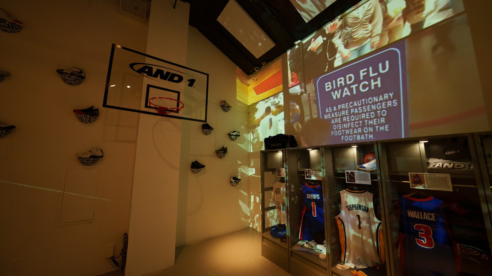Glass lockers holding basketball jerseys beneath a projected Bird Flu Watch sign, sneakers and an AND1 hoop on the wall