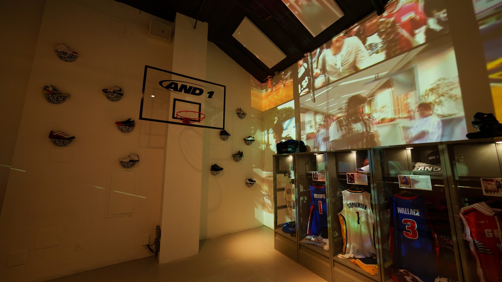 Low angle on an AND1 backboard and wall mounted sneakers beside glowing jersey lockers under projected footage
