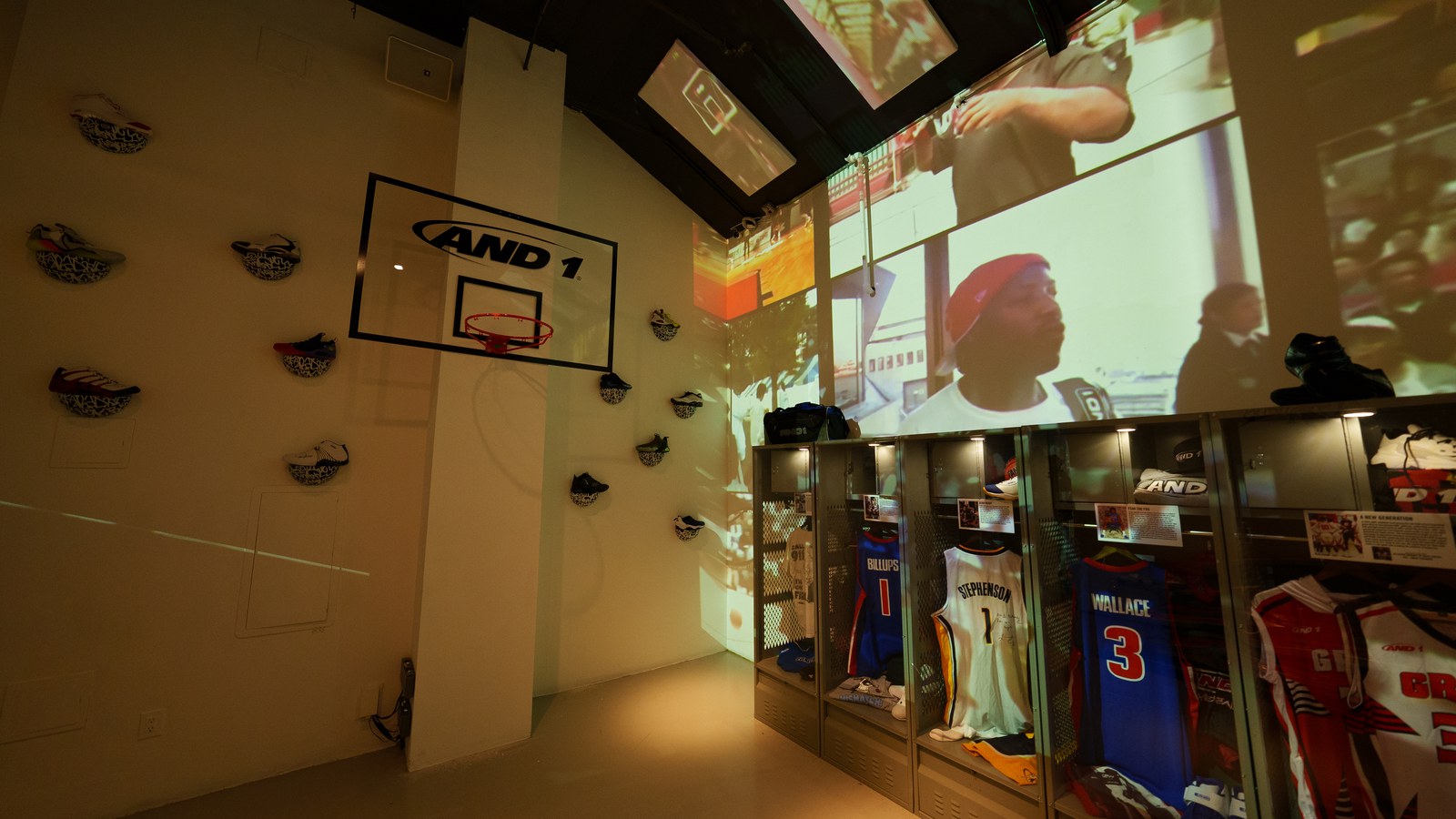 AND1 backboard and mounted sneakers on a wall beside lit lockers displaying jerseys under projected archive footage