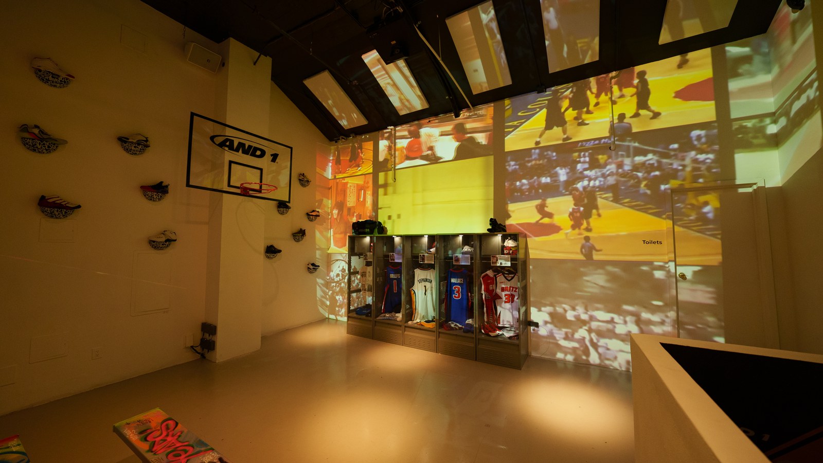 Wide view of a yellow lit room with jersey lockers, wall mounted sneakers and projected basketball game footage