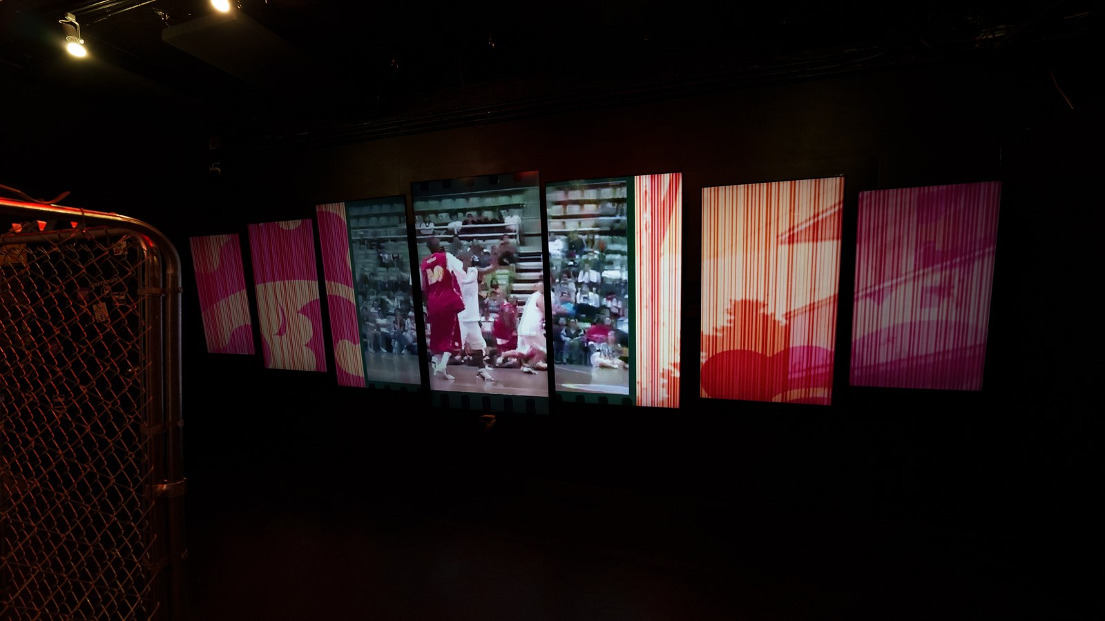 Dark room with a row of glowing panels showing red and pink basketball footage beside a chain link fence