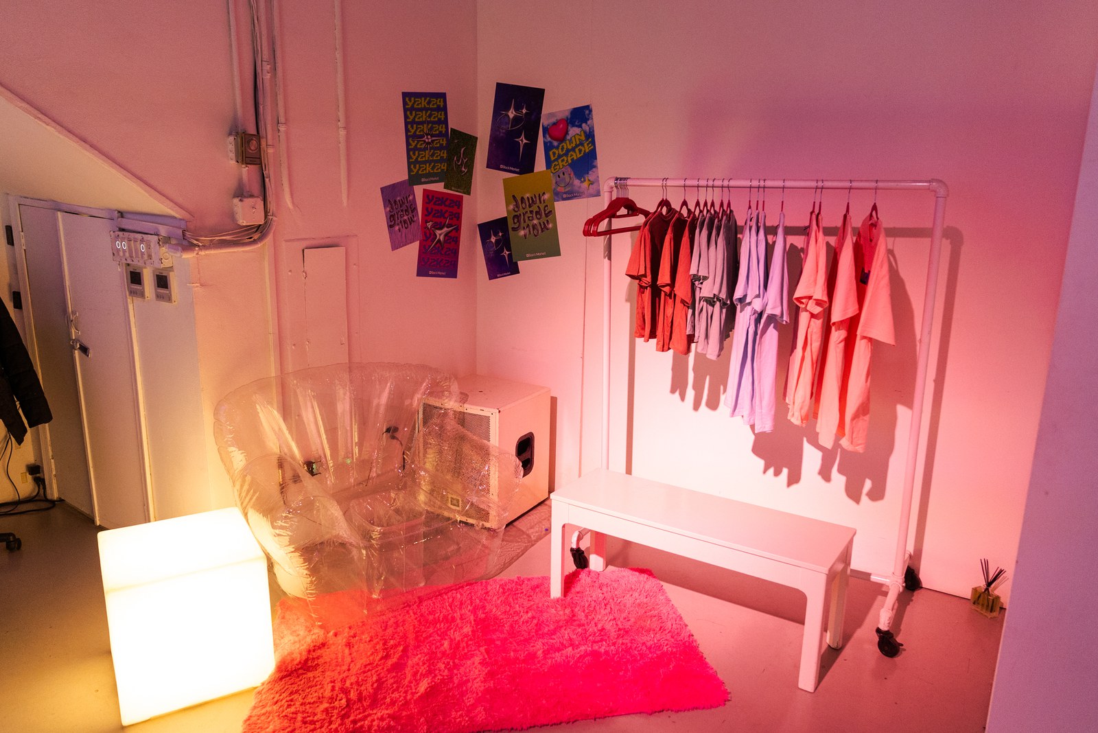 Small pink lit room with a clothes rack, inflatable chair, glowing cube lamp and a fuzzy red rug
