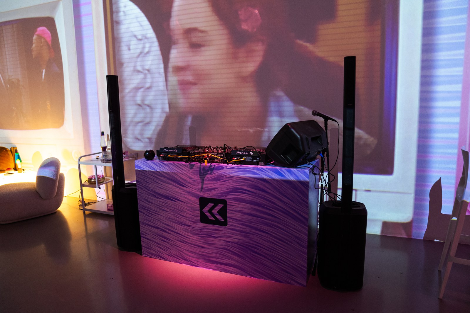 DJ booth draped in purple fabric between two tall speakers, projected face on the wall behind