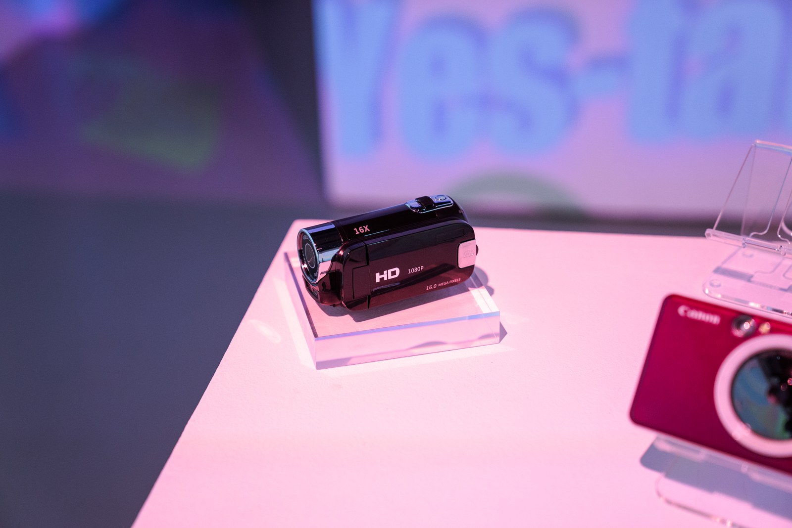 Small black HD camcorder on a clear acrylic stand under pink light beside a red instant camera