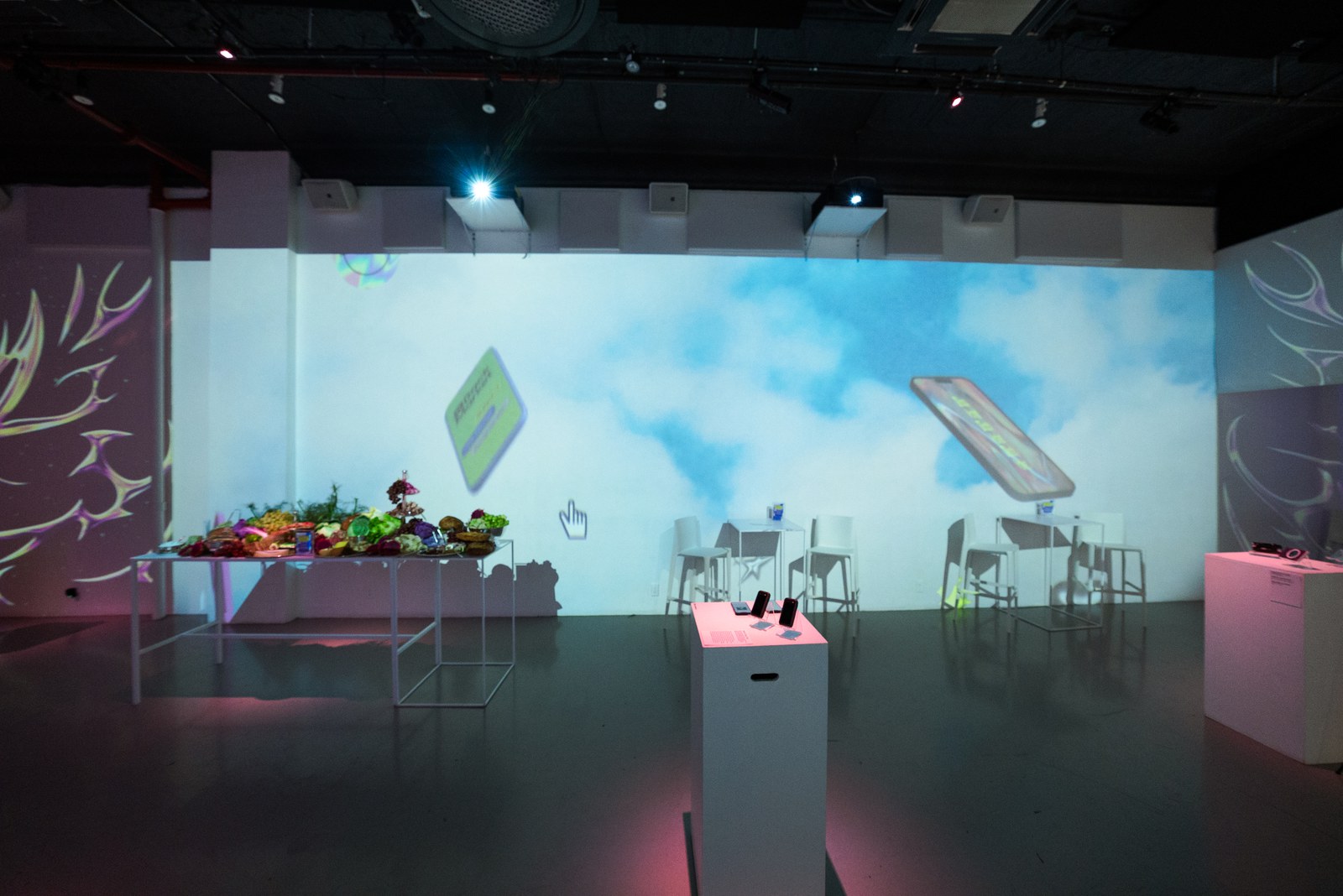 Wide room with white bar stools and projected cloud sky, phones on plinths, table of fresh produce on the left