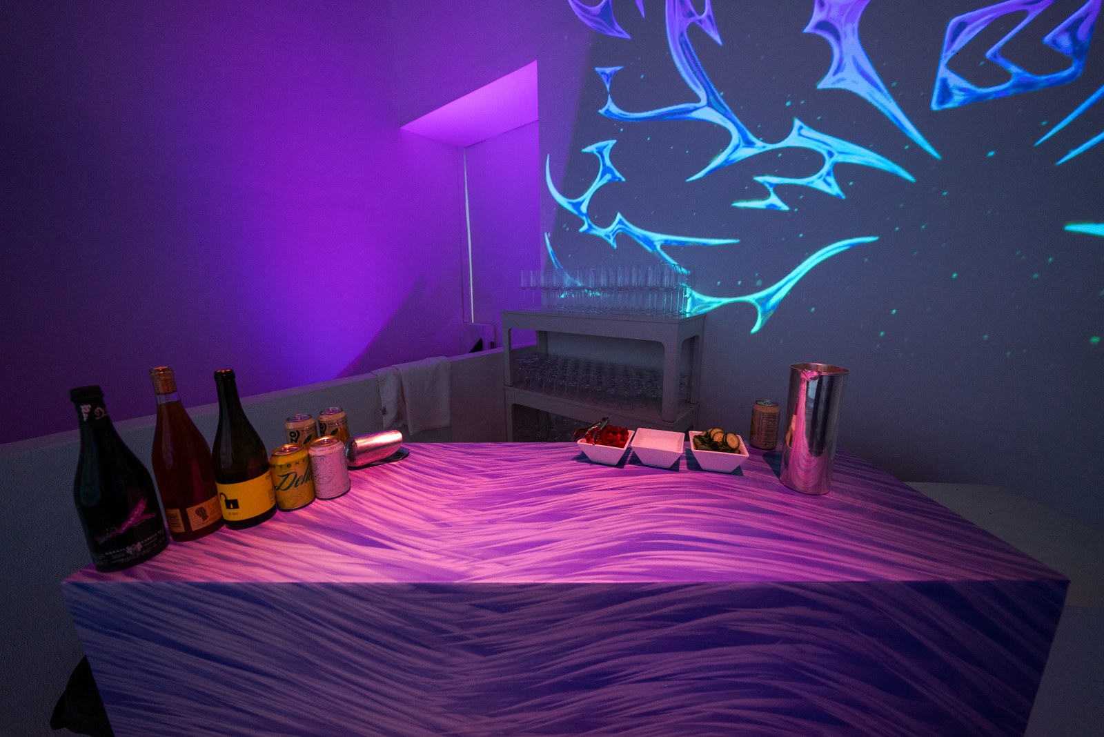 Bottles and snack bowls on a purple lit bar counter beneath a glowing blue projected pattern