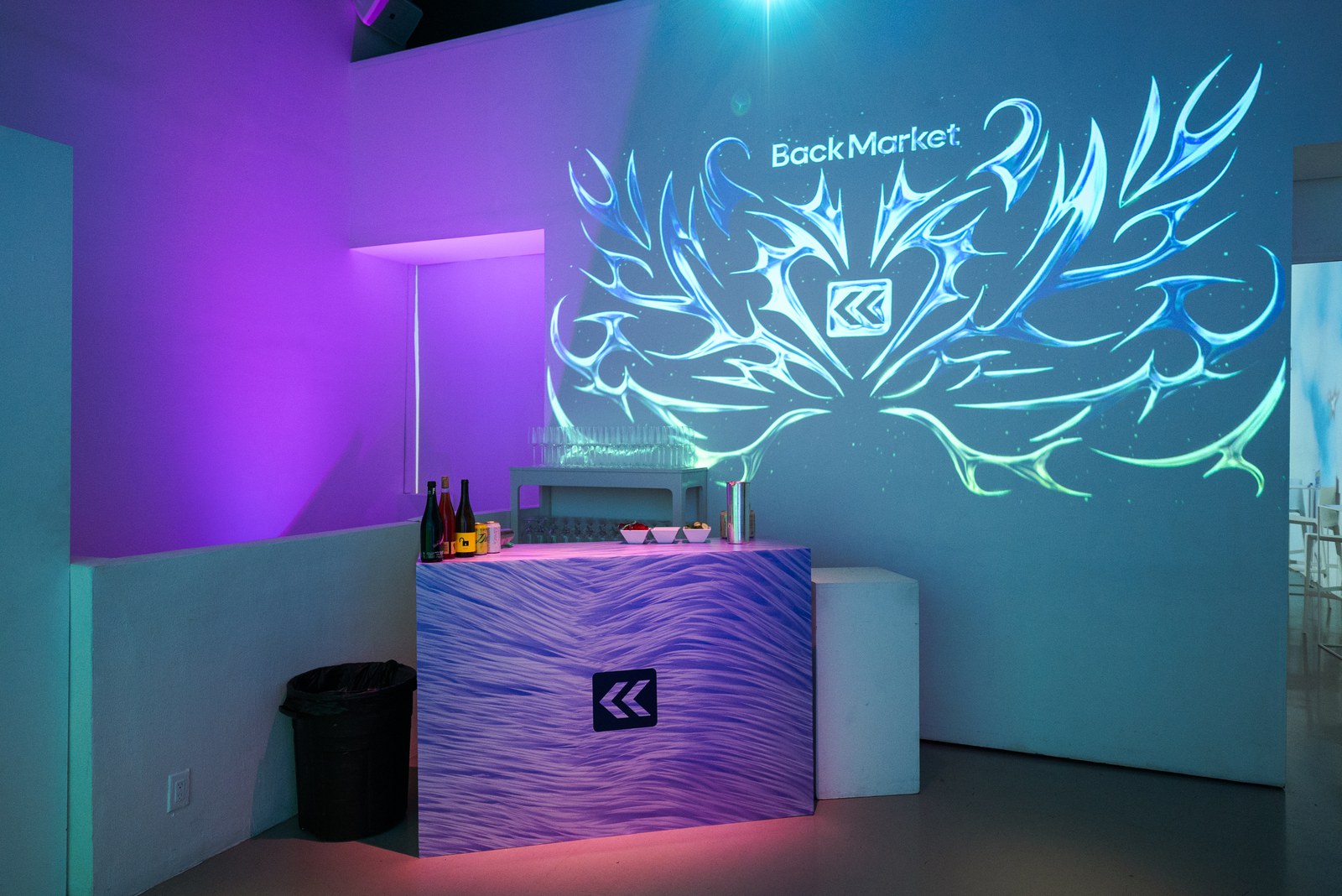 Purple lit bar with a wavy blue counter and bottles, Back Market logo projected above in swirling teal ornament
