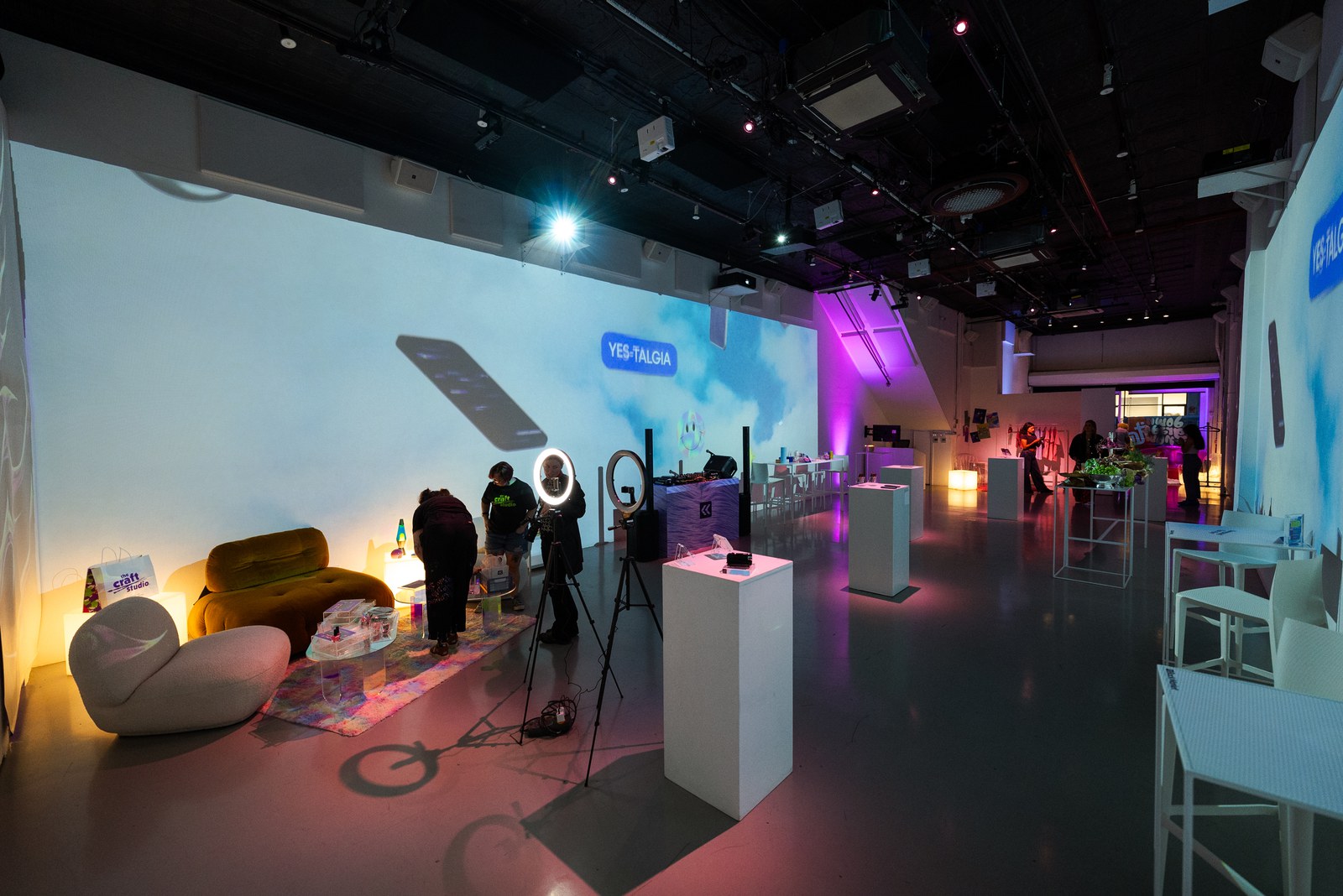 Long room with white display plinths, a ring light, a yellow sofa and a wall projection of clouds