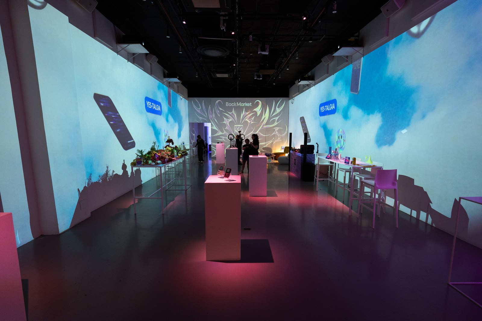 Long dark room with projected cloud sky walls, white plinths, pink chairs and guests at a far bar