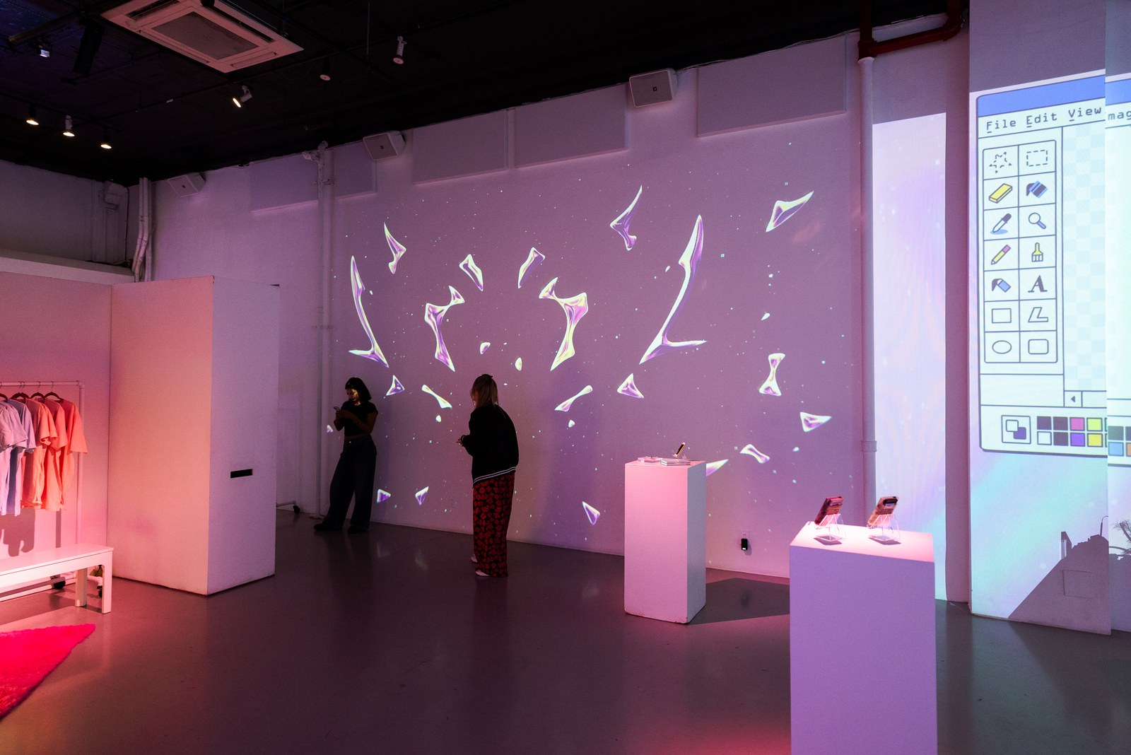 Two guests look at a wall of scattered projected shapes beside a paint program window and a clothing rack