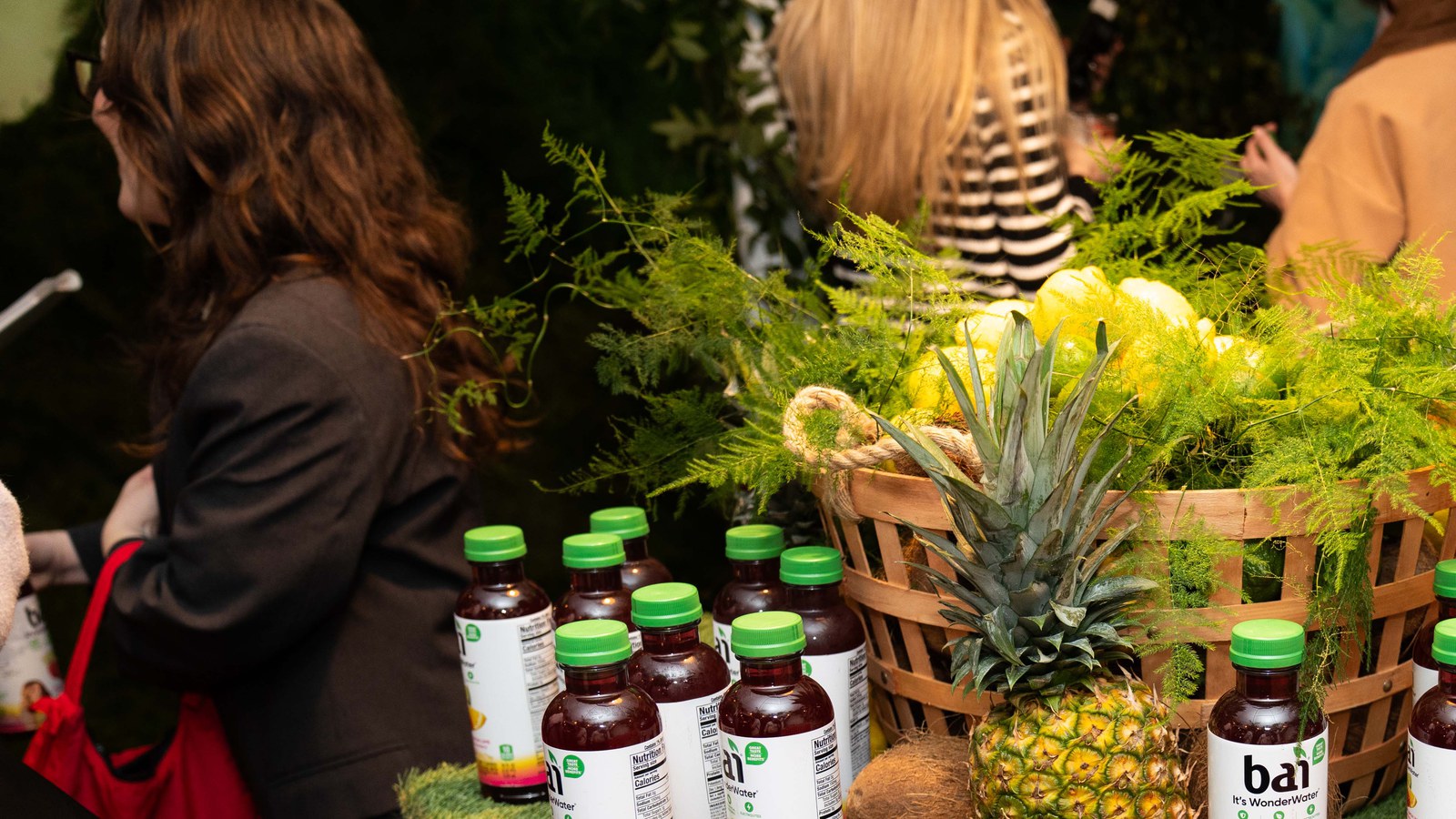 Green capped bottles of Bai juice sit beside a basket of pineapples, lemons and ferns as guests pass behind