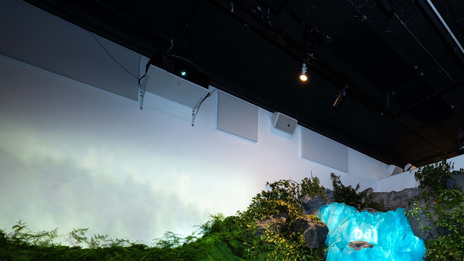 Ceiling rig with projector and lights above a white wall, green foliage and a blue lit crystal shape below