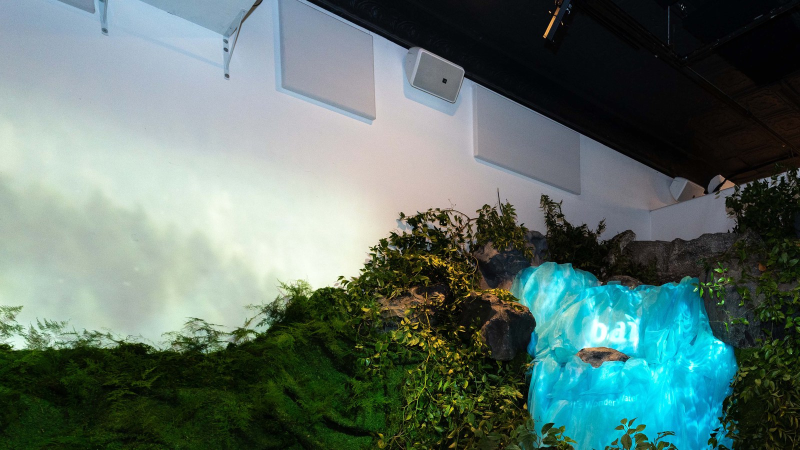 A glowing blue ice block bearing the Bai logo sits among greenery and rocks under a misty white wall