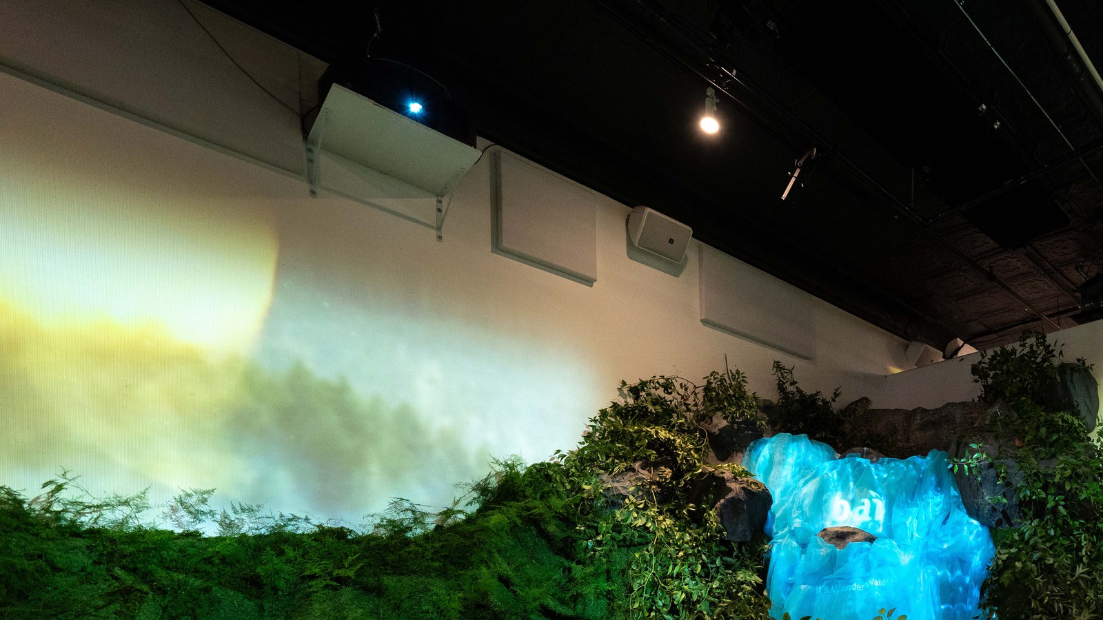 Glowing blue waterfall prop among green foliage under a white wall lit with warm projected light