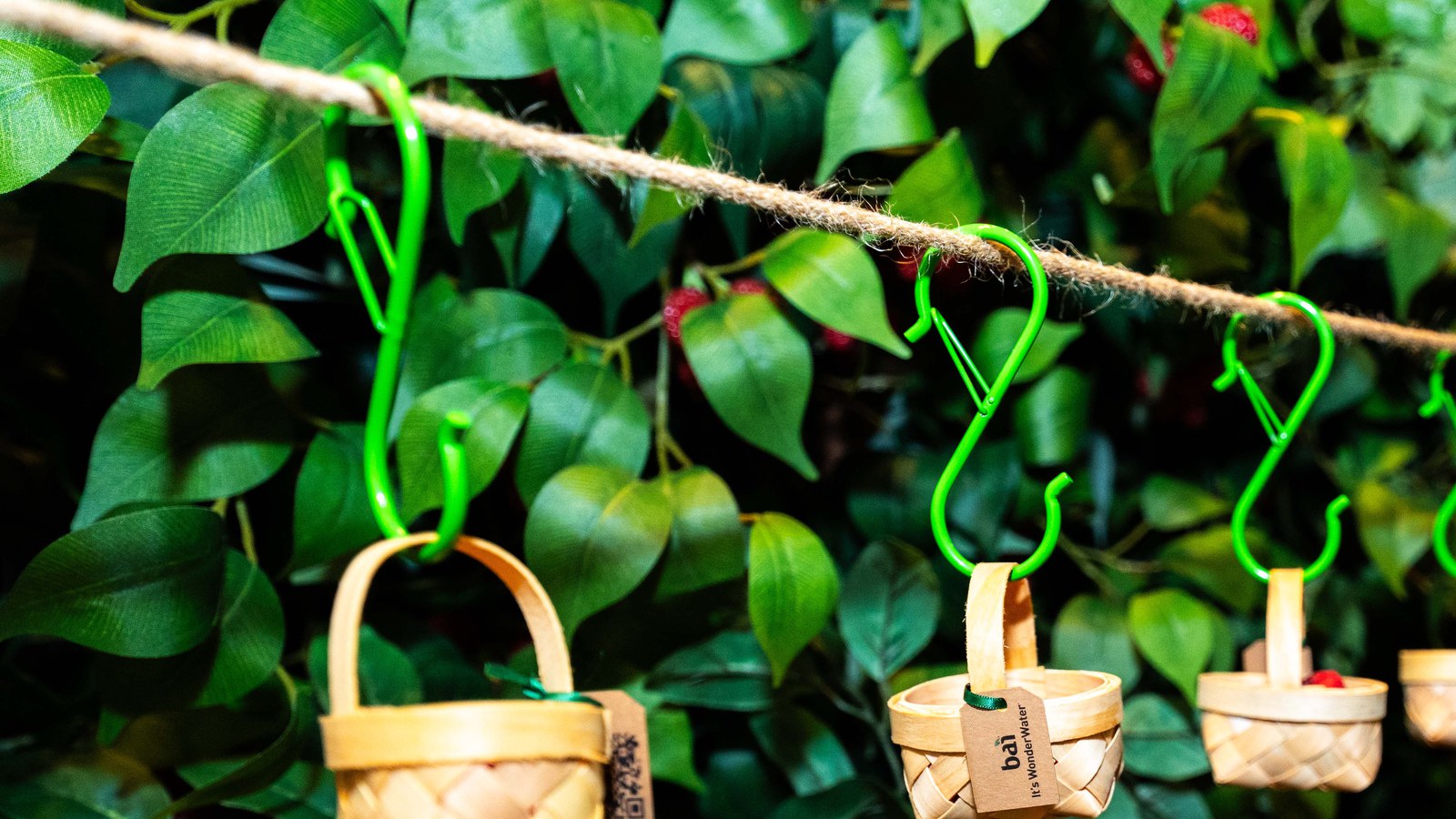 Small woven baskets hang from green hooks on a rope in front of dense green leaves