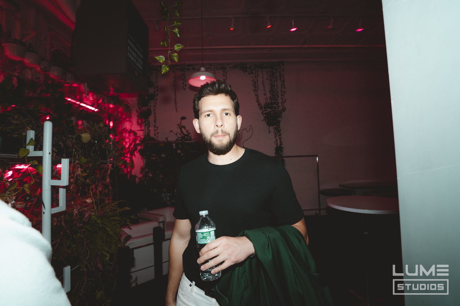 Man in a black tee holding a water bottle in a plant filled room lit red
