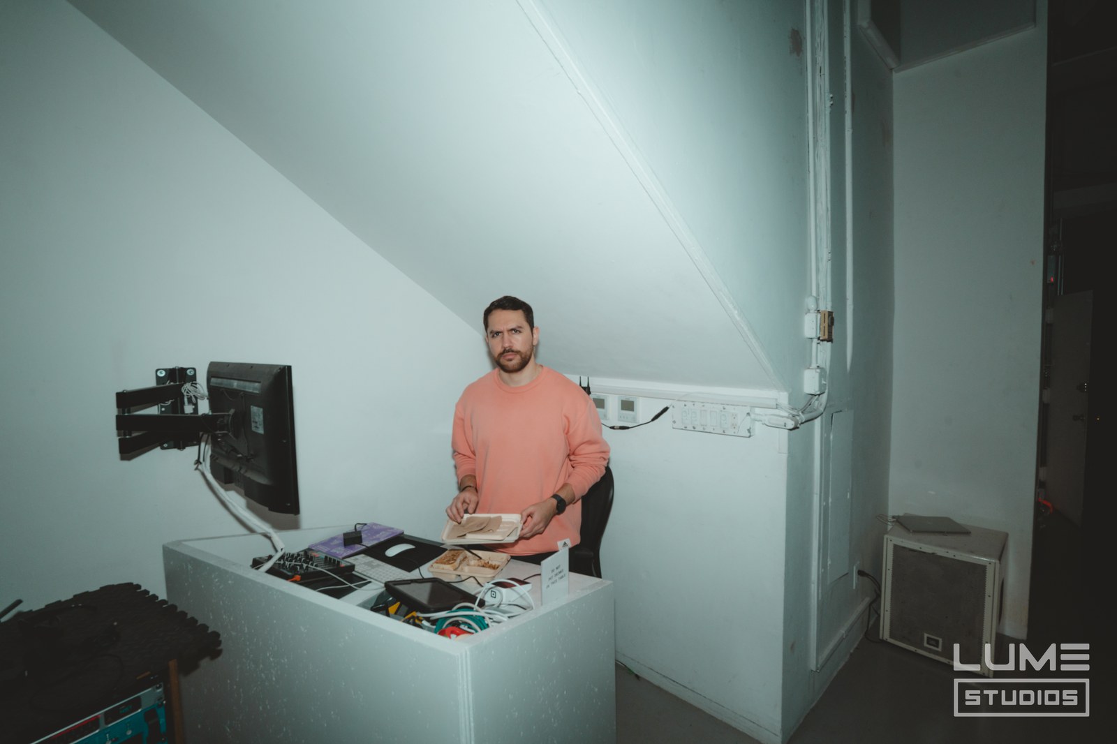 Man in a salmon sweater stands at a small workstation with a wall mounted monitor in a white stairwell corner
