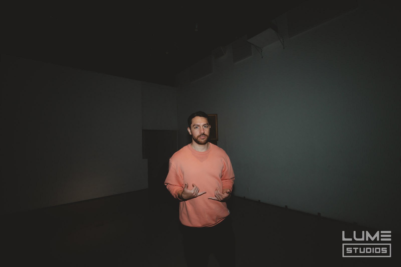 Man in a peach sweater gestures with both hands while speaking in a dimly lit room