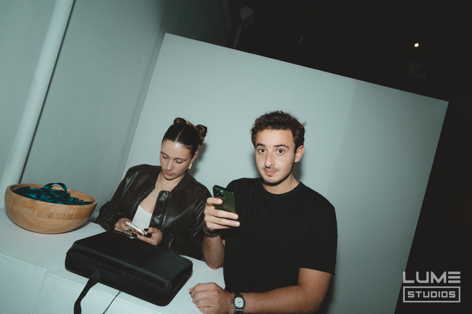 Two guests at a white table lit by flash, both holding phones, a wooden bowl beside them