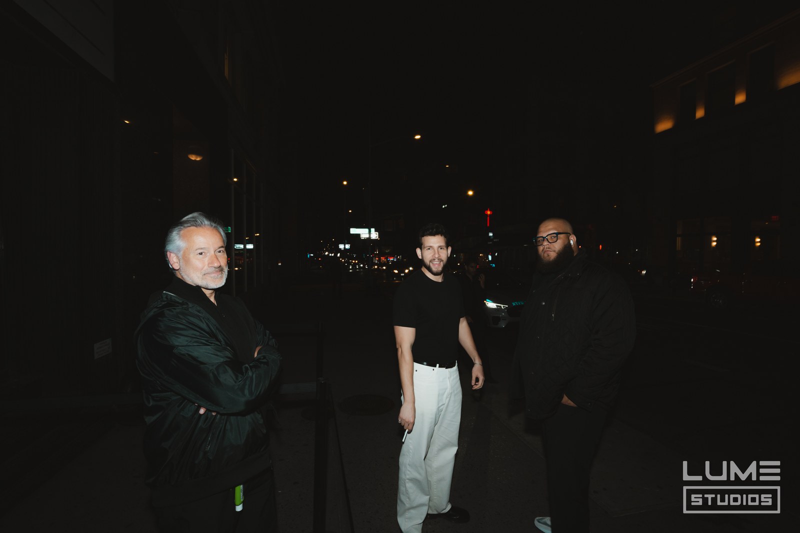 Three men stand on a dark city street at night with blurred traffic lights behind them