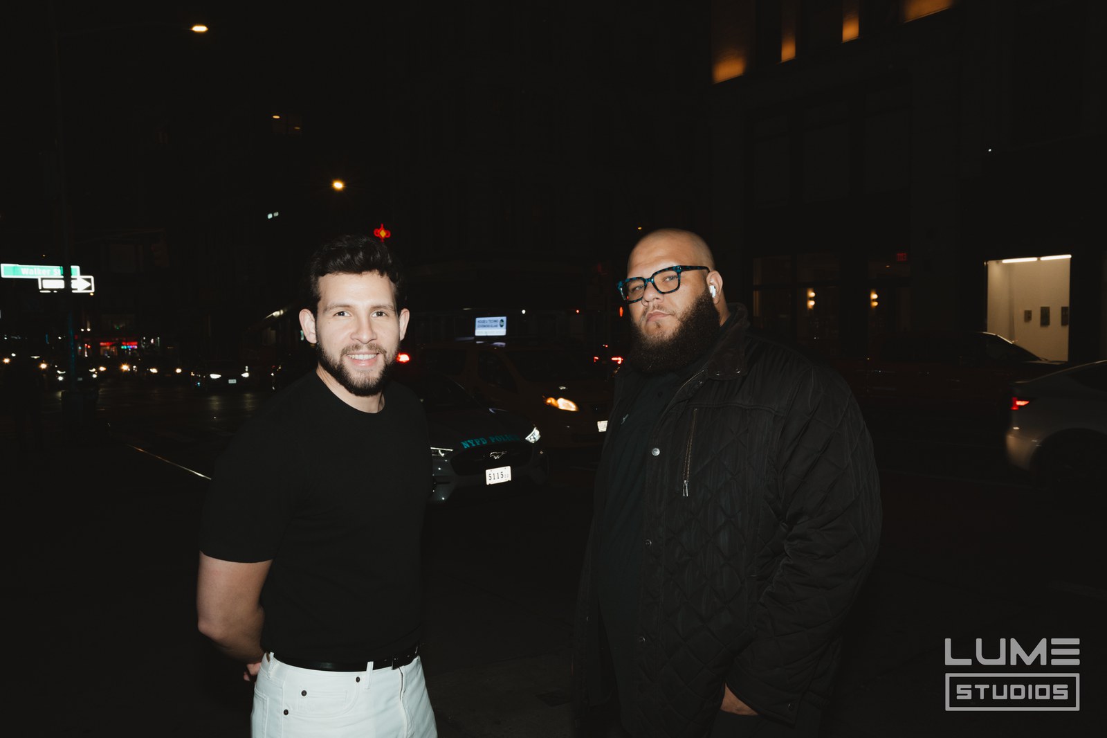 Two men stand on a city street at night with car headlights and streetlights behind them