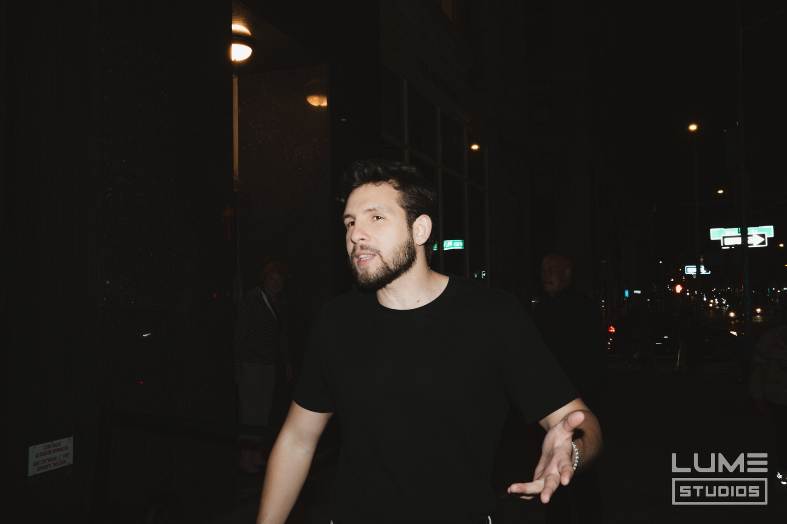 Man in a black tee gestures on a dark city street at night with traffic lights behind him