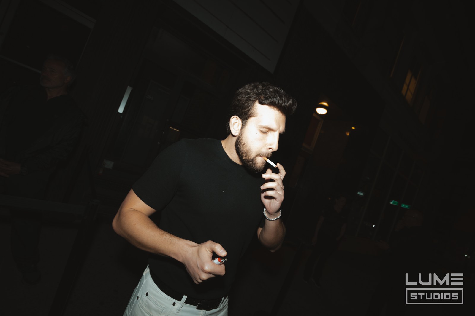 Man in a black tee lights a cigarette at night, lit by camera flash against a dark street