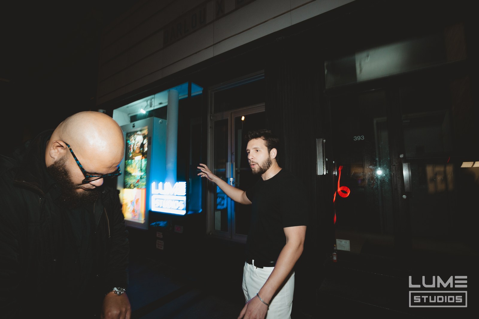 Two men talk on a night sidewalk outside a lit storefront, one gesturing mid sentence